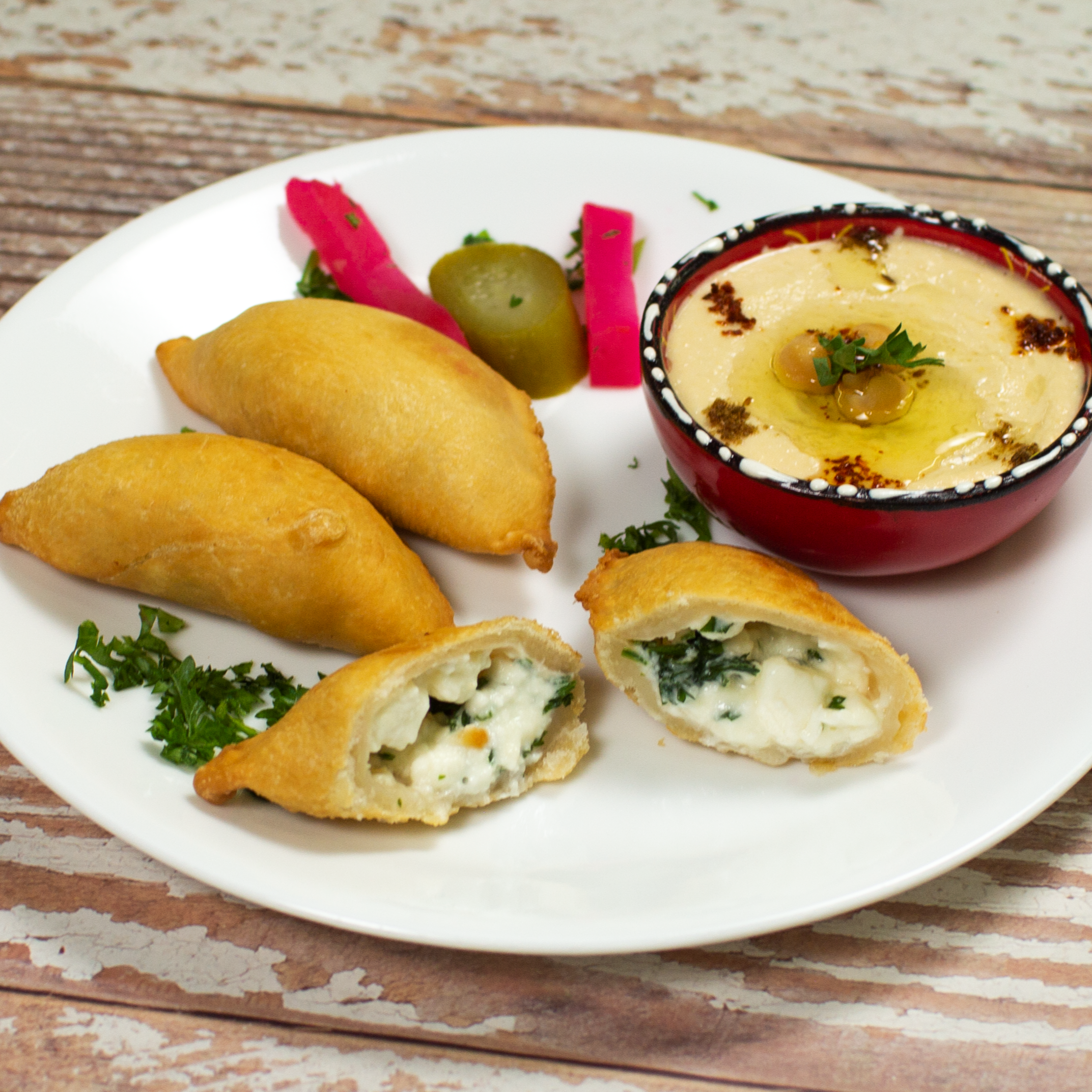 A plate featuring Tayybeh's Cheese Samboosak, deliciously crisp homemade spinach and cheese pastries, accompanied by a bowl of hummus with olives and spices, plus a side of pickled vegetables.