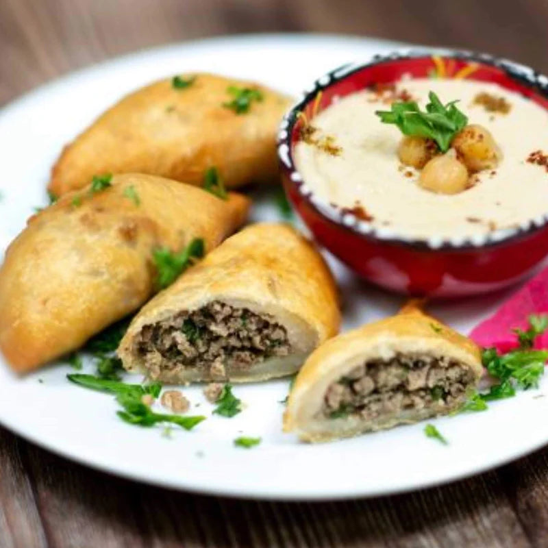 On a plate, four crispy pockets from Tayybeh's Frozen Beef Sambousak are filled with ground beef and garnished with parsley, sitting beside a bowl of hummus adorned with chickpeas and herbs, infused with traditional spices.