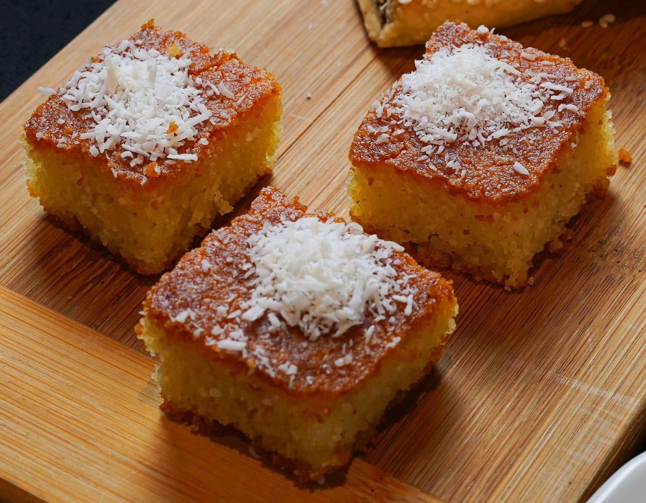 Three slices of Tayybeh's lightly sweetened semolina Coconut bars are generously topped with shredded coconut and elegantly arranged on a rustic wooden board.