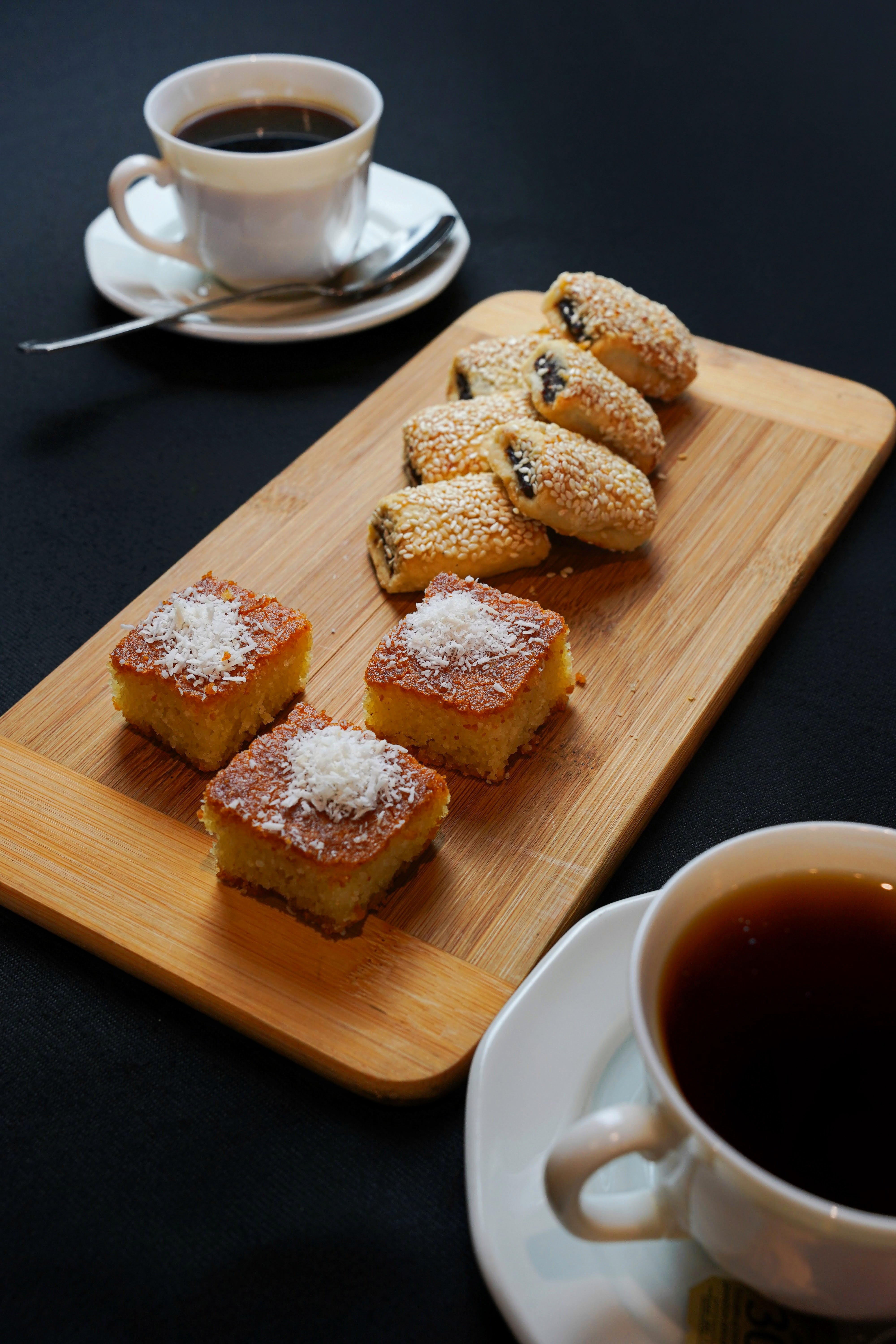 The "Coffee Break Package 2" by Tayybeh, featuring a wooden tray with two cups of fair-trade coffee, coconut-topped nammoura squares, and sesame-seed-covered barazek pastries, rests elegantly on a black table.