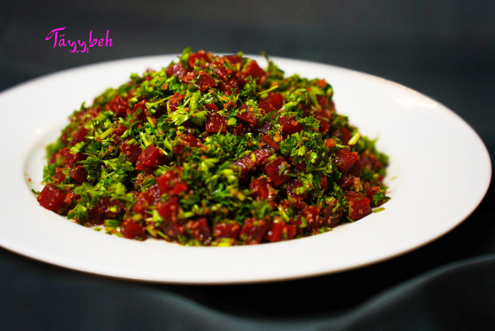 A plate of Tayybeh's Beet Tabbouleh featuring finely chopped parsley, diced red vegetables, and a touch of pomegranate molasses set against a dark background.