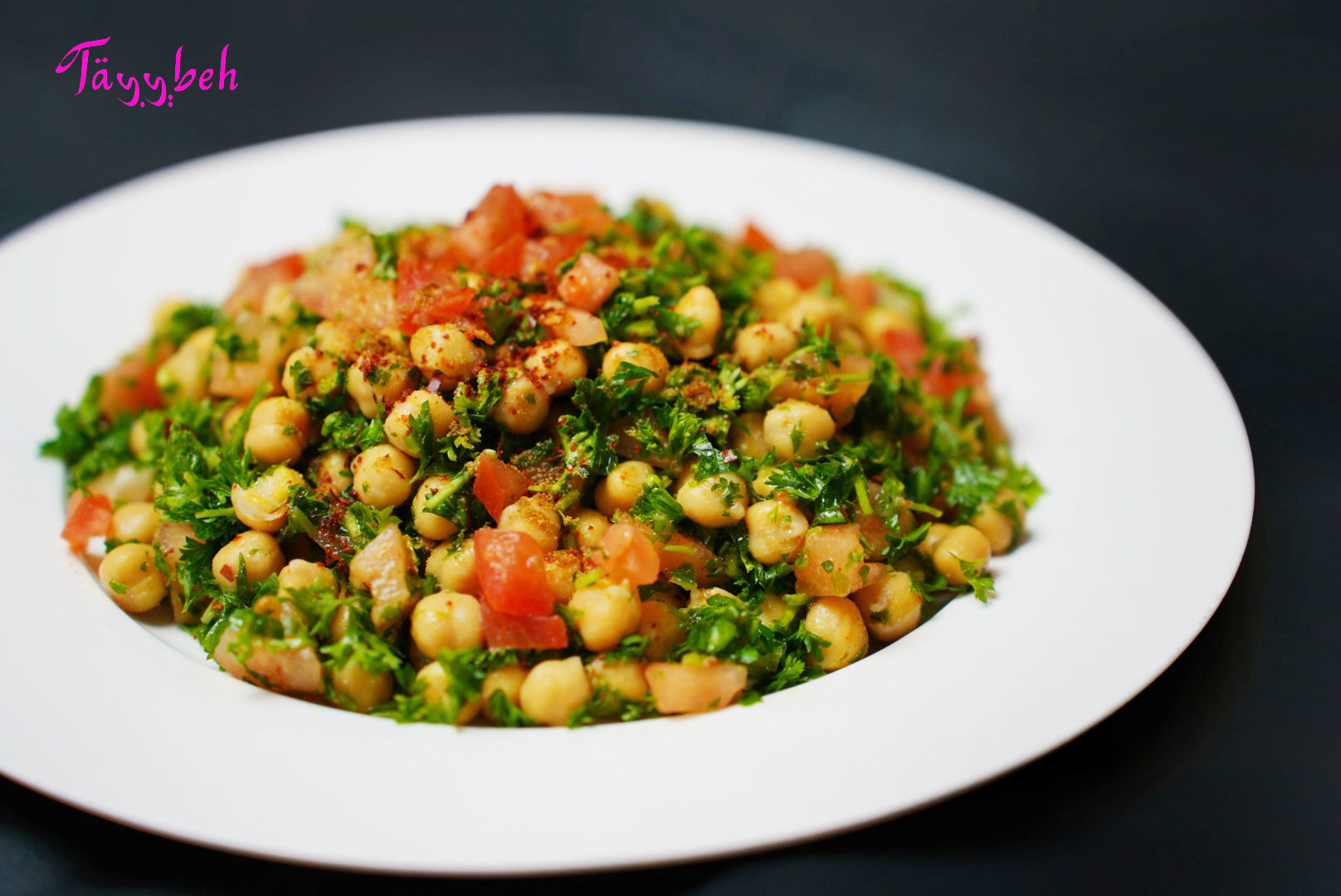 Tayybeh's Chickpea Salad features a blend of chickpeas, diced tomatoes, and chopped parsley, offering numerous health benefits, and is topped off with a sprinkle of spices.