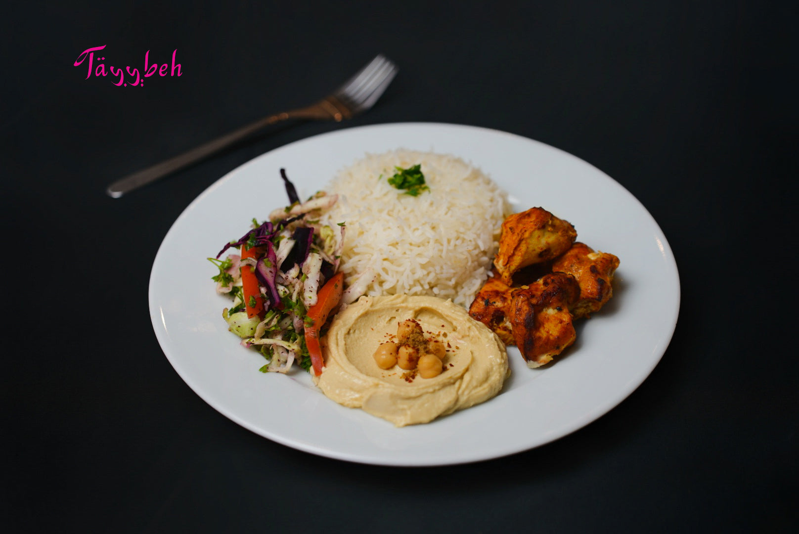 A plate of Tayybeh's Sheesh Tawook Meal, featuring fragrant Basmati rice with herbs, grilled chicken skewers, creamy hummus dip garnished with chickpeas, and a side salad, sits on a dark background with a fork.