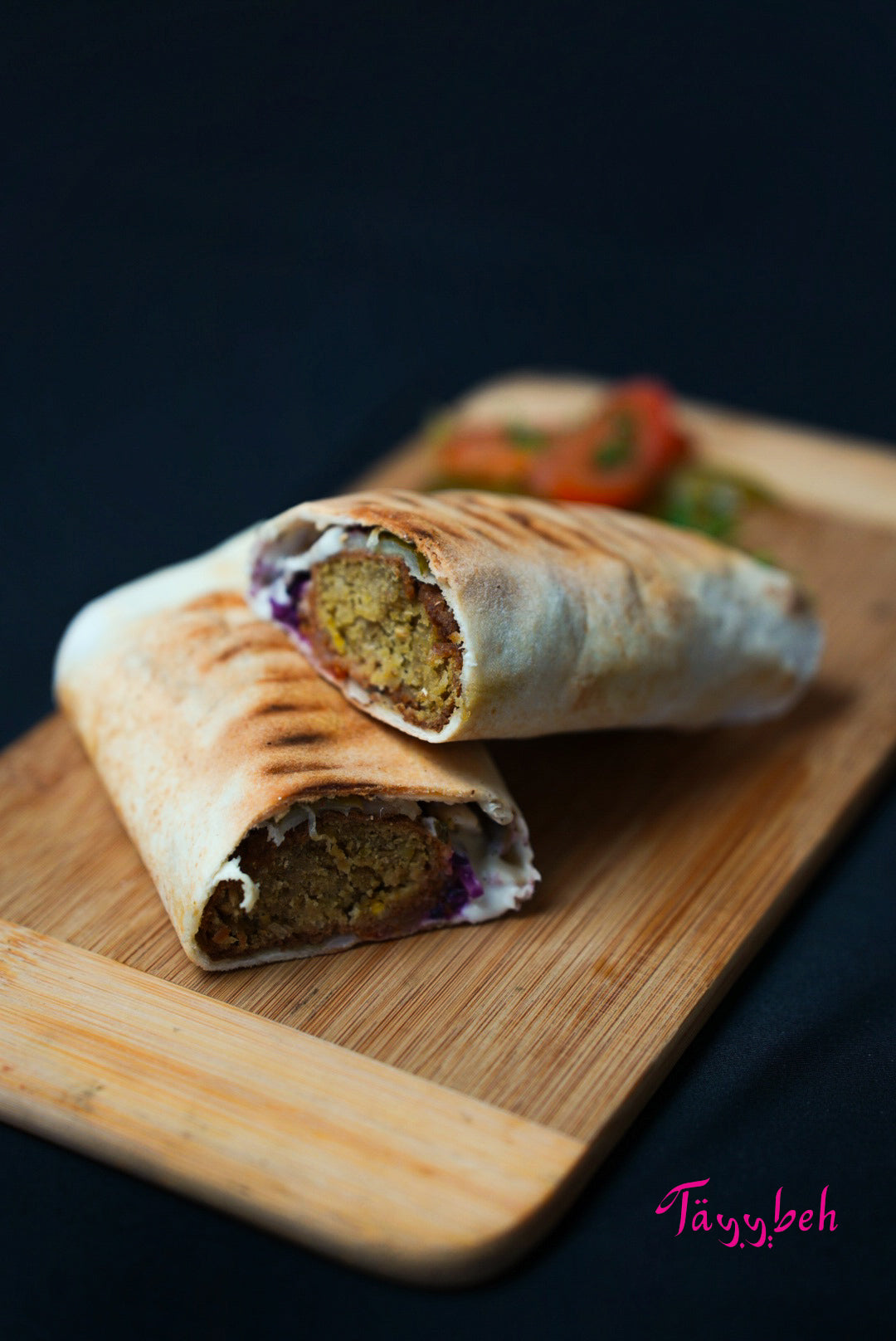 A Tayybeh Falafel Wrap, sliced in half and topped with tahini sauce, sits on a wooden board with blurred tomatoes and greens in the background.