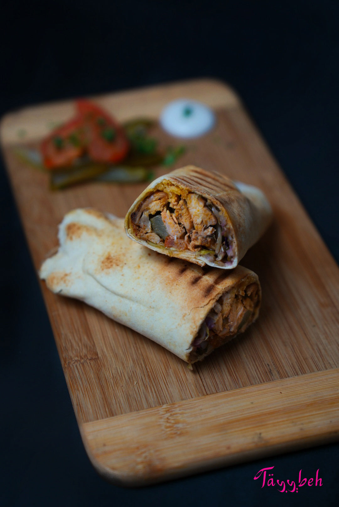 A tantalizing Chicken Shawerma wrap from Tayybeh, nestled on a wooden board, is paired with crisp pickles, juicy tomato slices, and a dollop of creamy garlic sauce.