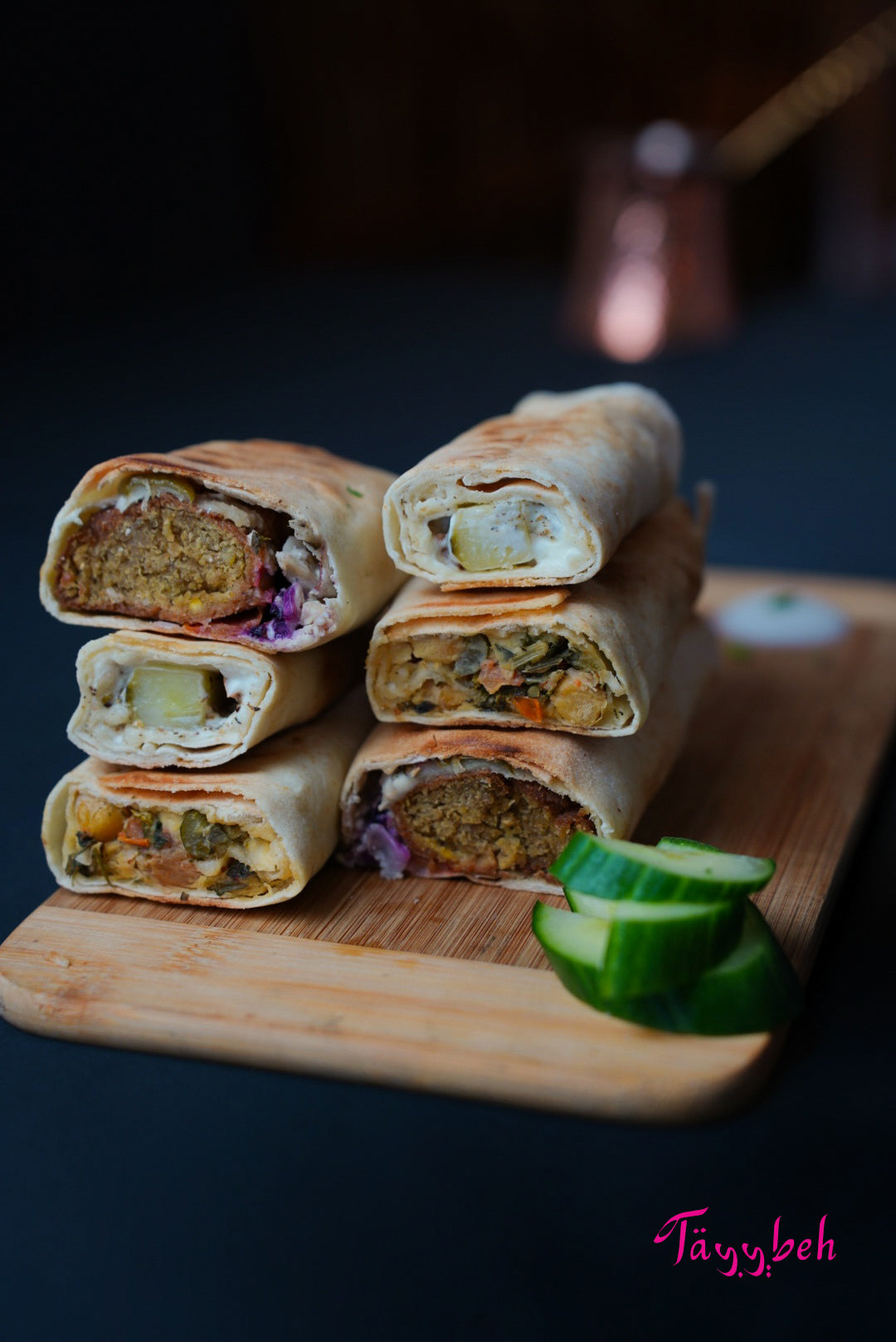 A platter of Assorted vegetarian Wraps by Tayybeh, filled with vibrant vegetables, is displayed on a wooden board alongside sliced cucumbers.