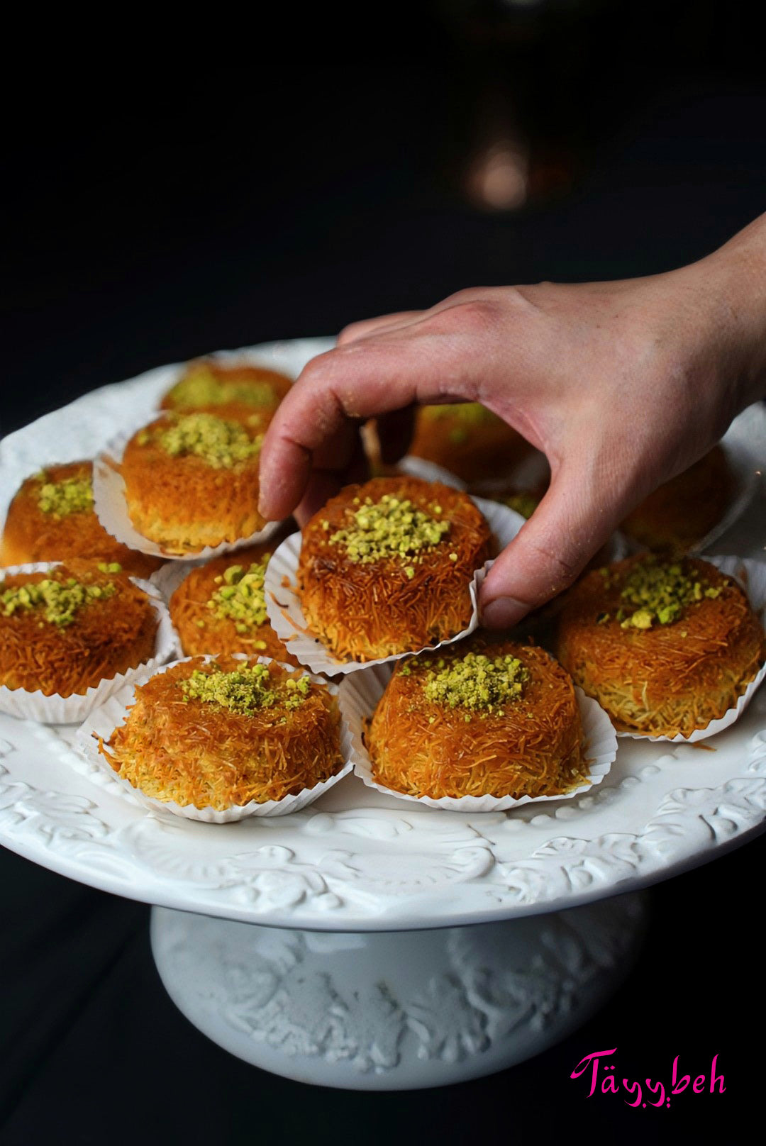 A hand reaches for a Knafeh, a cream-filled dessert topped with pistachios and made by Tayybeh, displayed on a white cake stand amidst similar Middle Eastern treats.