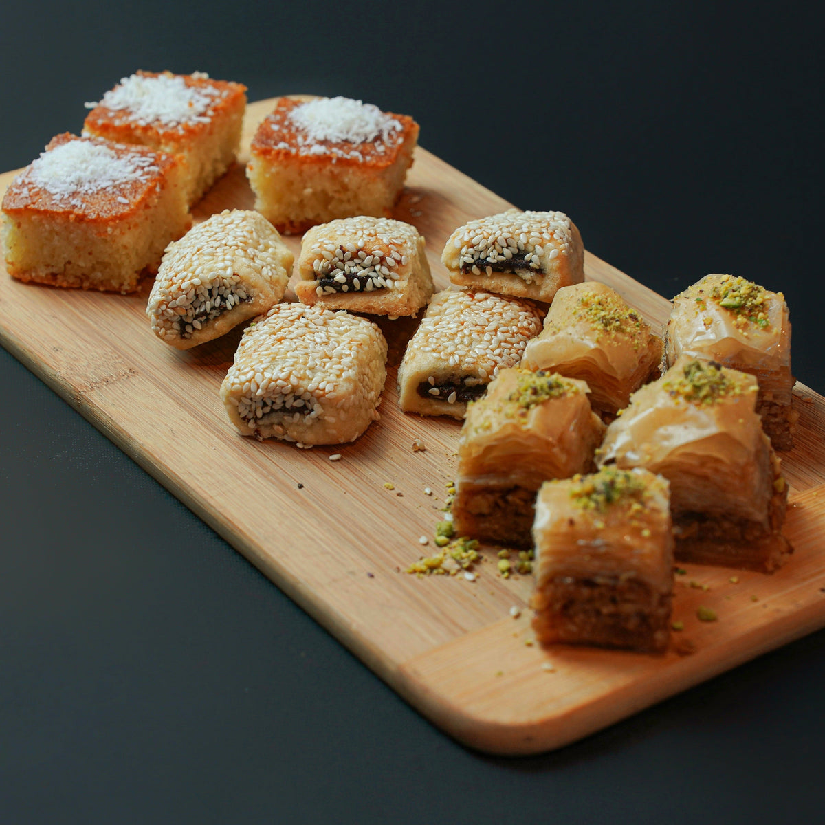 A wooden board featuring the Assorted Dessert Platter by Tayybeh, which includes coconut-topped Nammoura squares, sesame-covered pastry rolls, and pistachio-topped baklava.