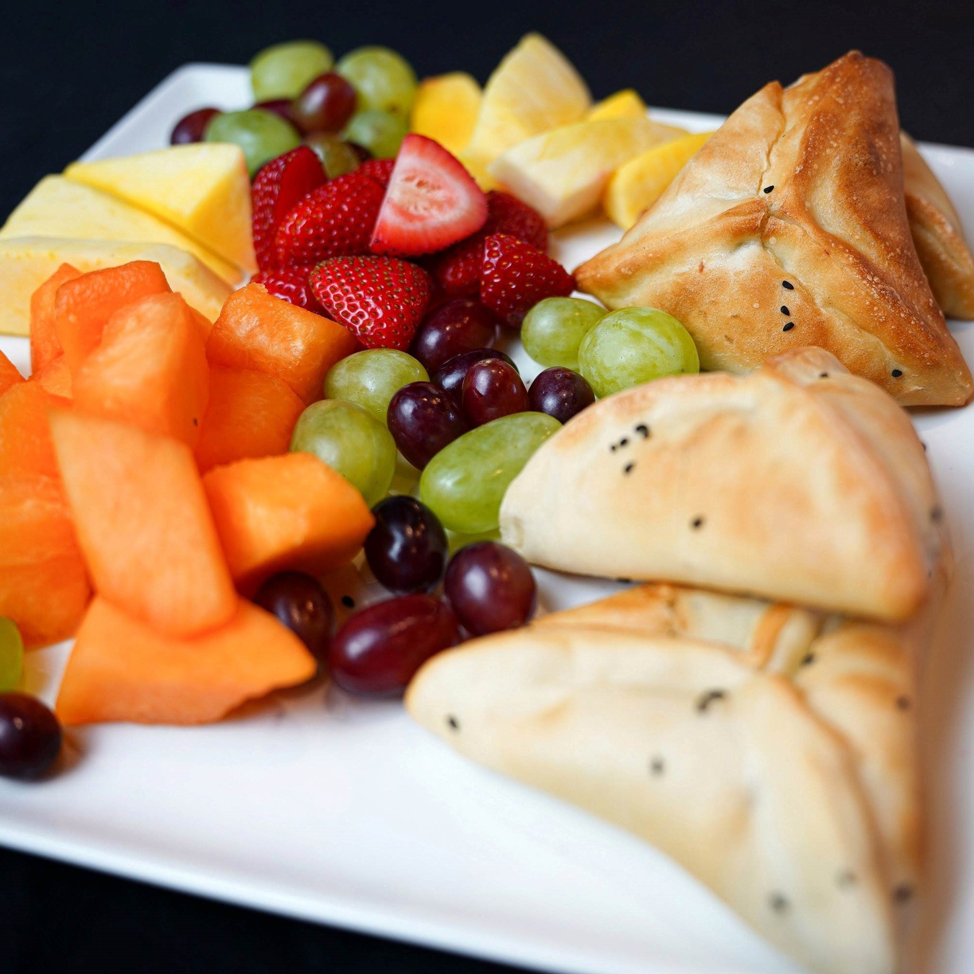 The Light Breakfast by Tayybeh showcases a beautifully arranged platter of assorted fresh fruits, including strawberries, melon, grapes, and pineapple. This is accompanied by delectable stuffed pastries topped with seeds and delightful cheese pockets.