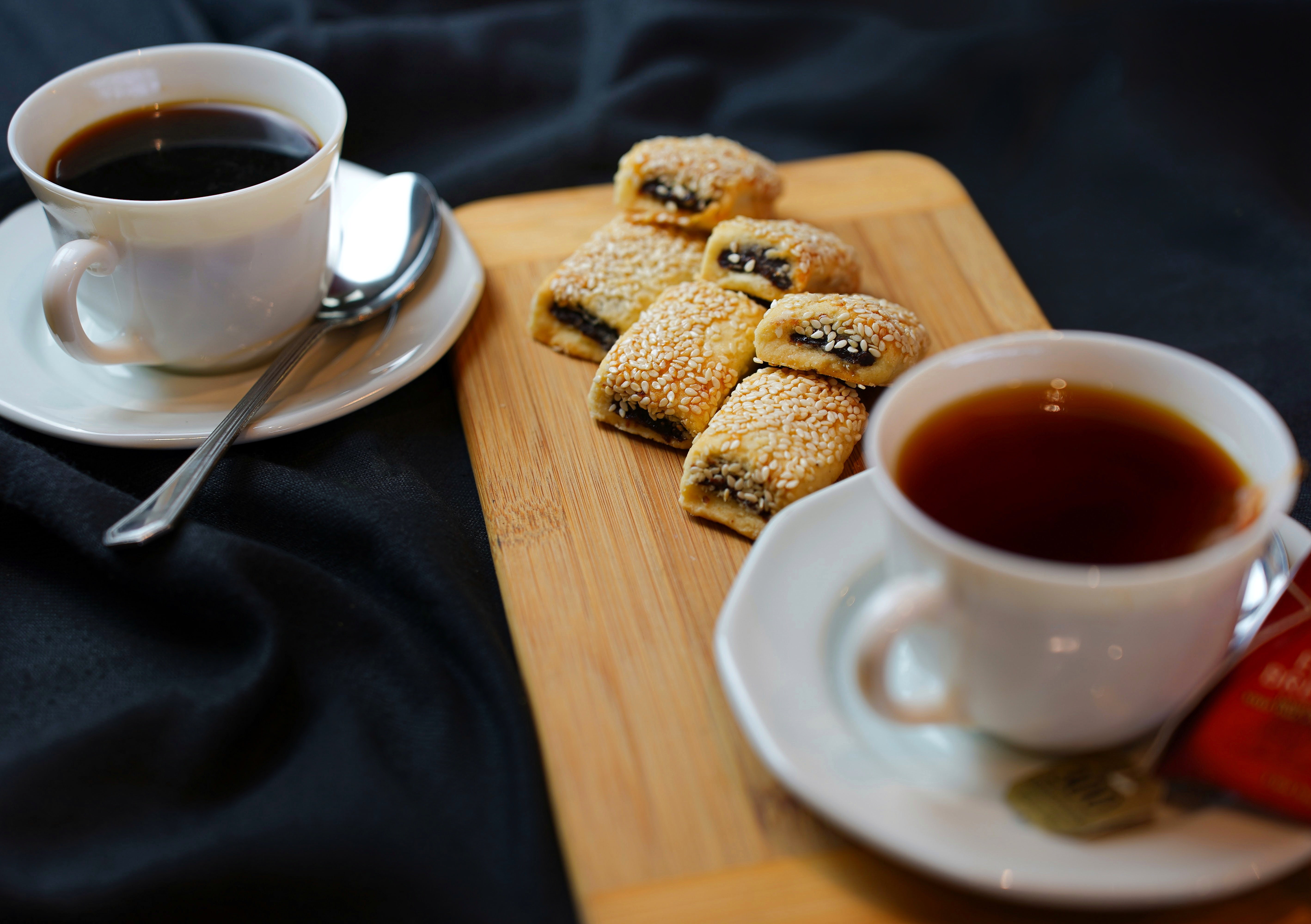 Tayybeh's Coffee Break Package 1 includes two cups of tea on saucers with spoons, accompanied by a wooden board featuring date cookies and sesame pastries, all elegantly arranged on a black tablecloth.