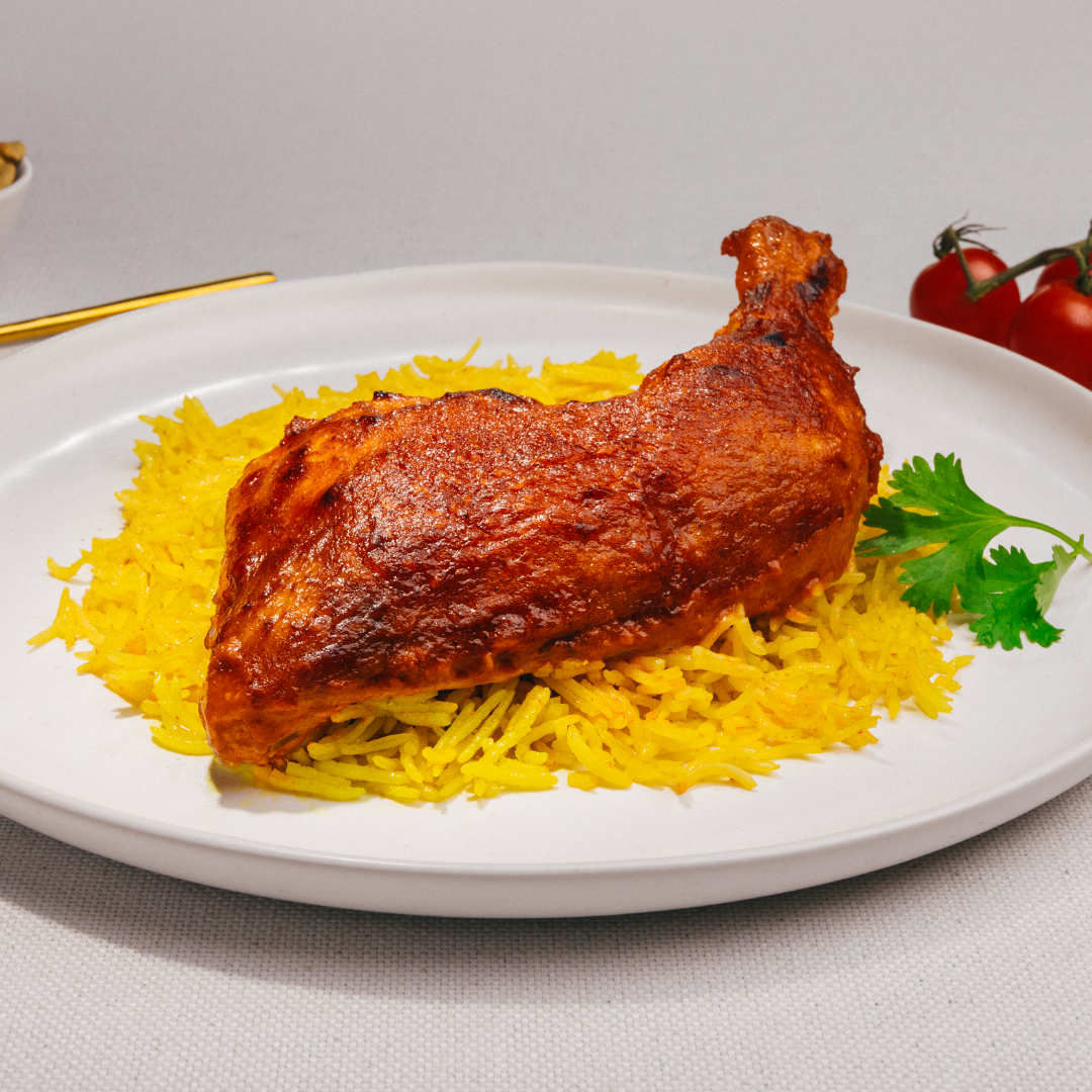 The Mesahhab Roast Chicken Single Frozen Meal by Tayybeh features perfectly roasted chicken on a bed of spiced basmati rice, garnished with parsley, elegantly presented on a white plate. In the background, tomatoes and a fork enhance this gluten-free delight.