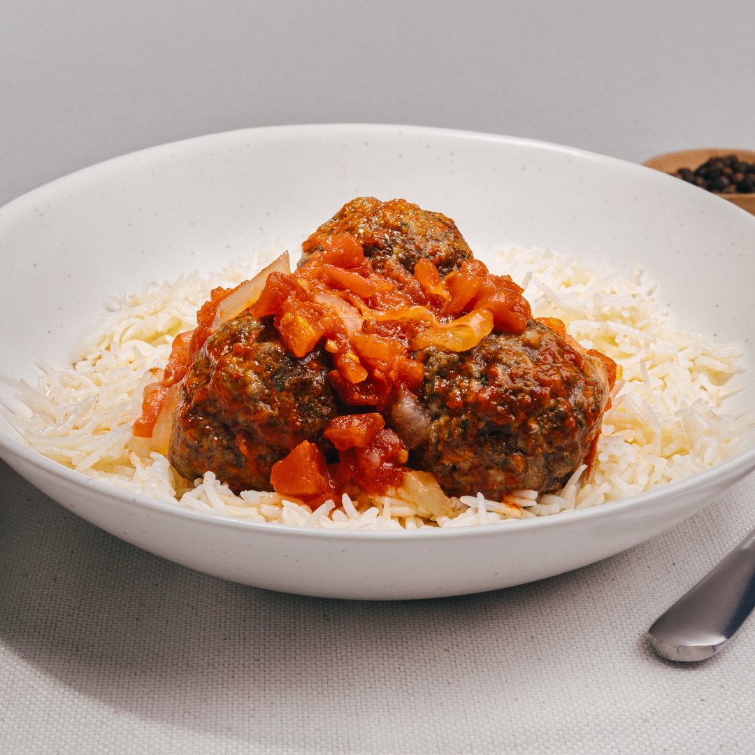 A white bowl features the "Meatball with Tomato Sauce Single Frozen Meal" by Tayybeh, showcasing two large beef meatballs in tomato sauce, garnished with chopped tomatoes and onions. This gluten-free dish is a prime example of ready-to-eat meals. A fork is partially visible on the right side.