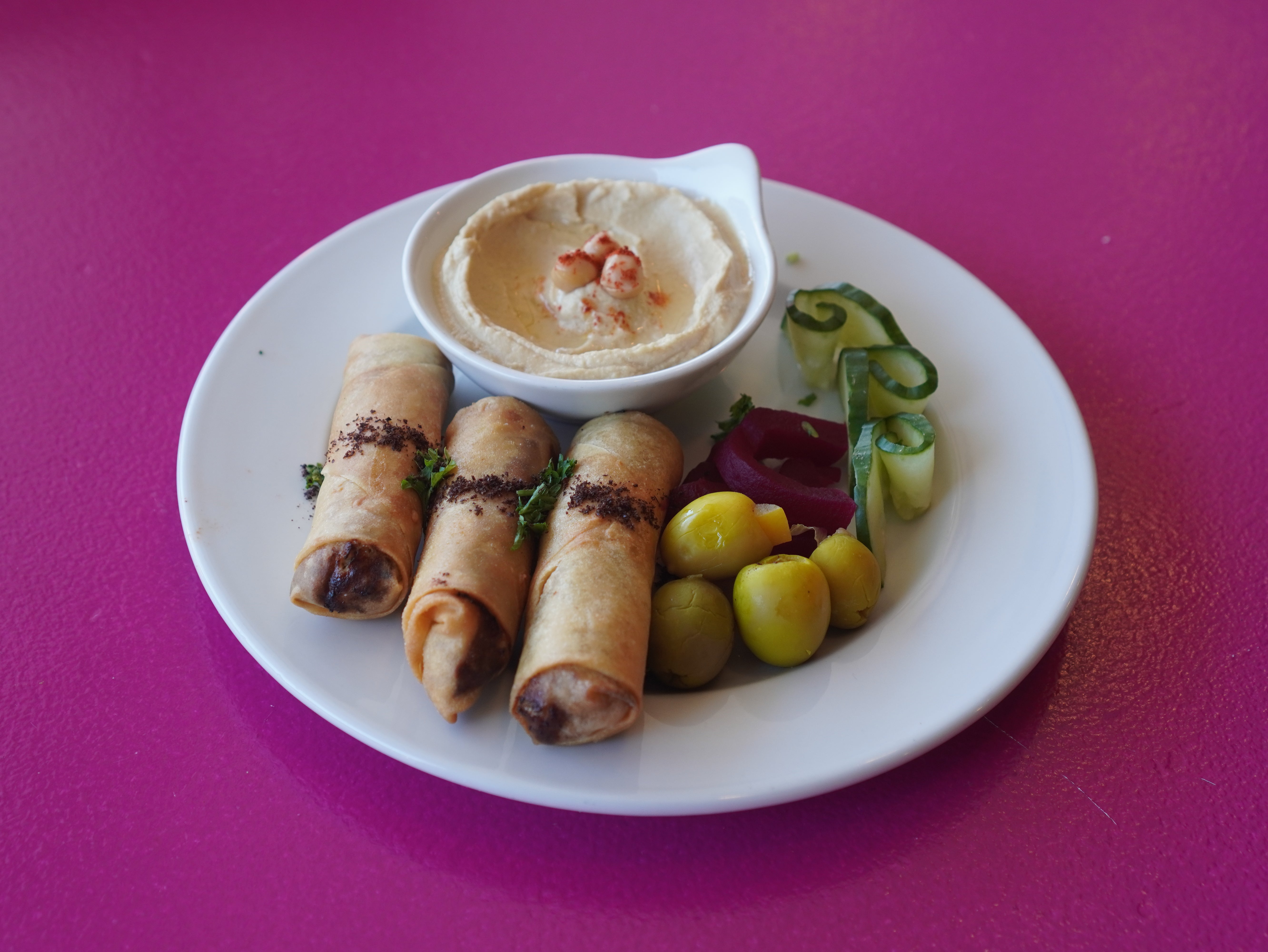 A plate featuring three crispy golden Musakhan Chicken Rolls by Tayybeh, accompanied by a dish of hummus sprinkled with paprika and spices, alongside side garnishes of cucumber, pickled vegetables, and herbs on a pink surface.