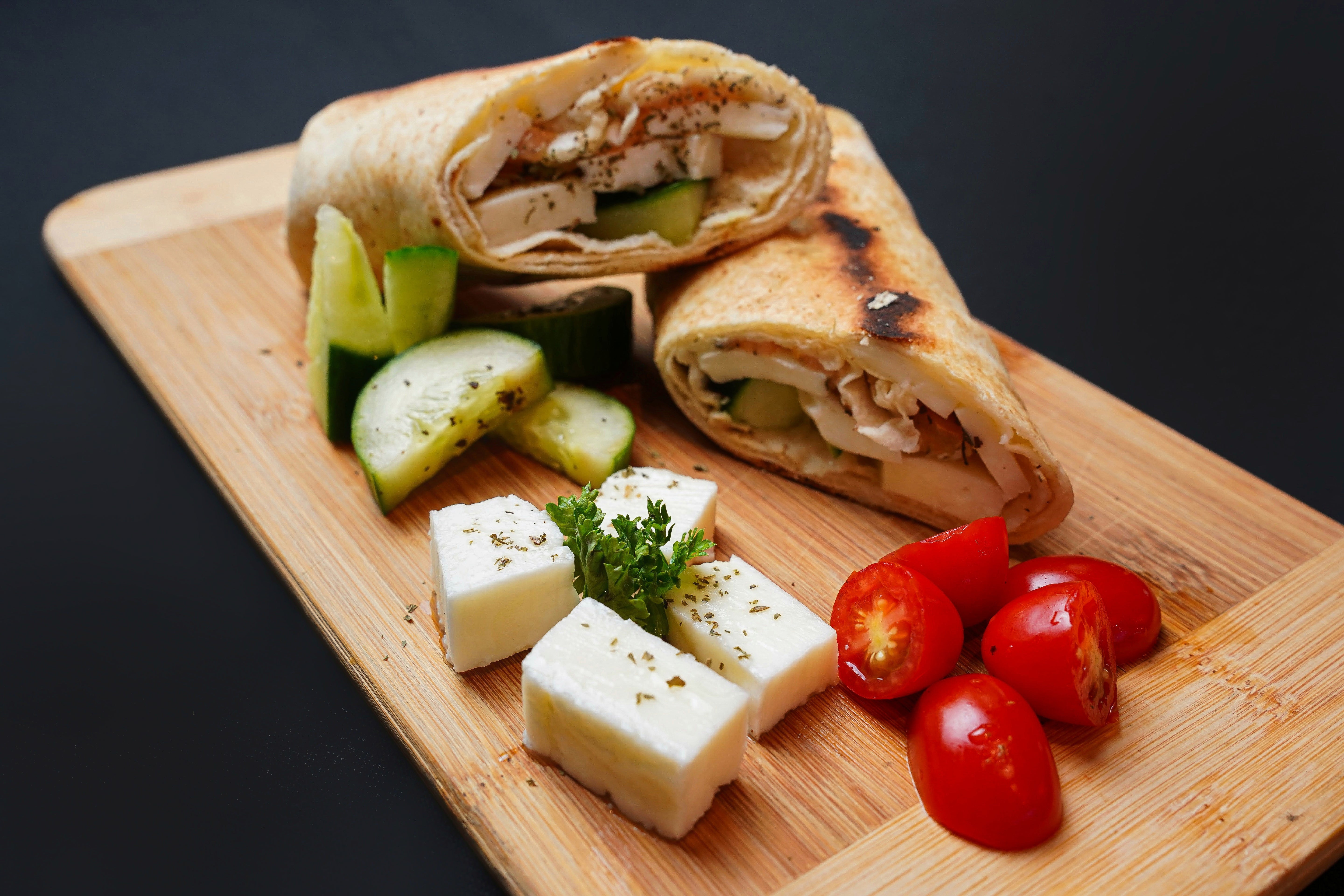 Tayybeh's Halloumi Wrap is served sliced on a wooden board, filled with chicken and vegetables, and garnished with cucumber slices, cherry tomatoes, grilled halloumi, and a hint of oregano.
