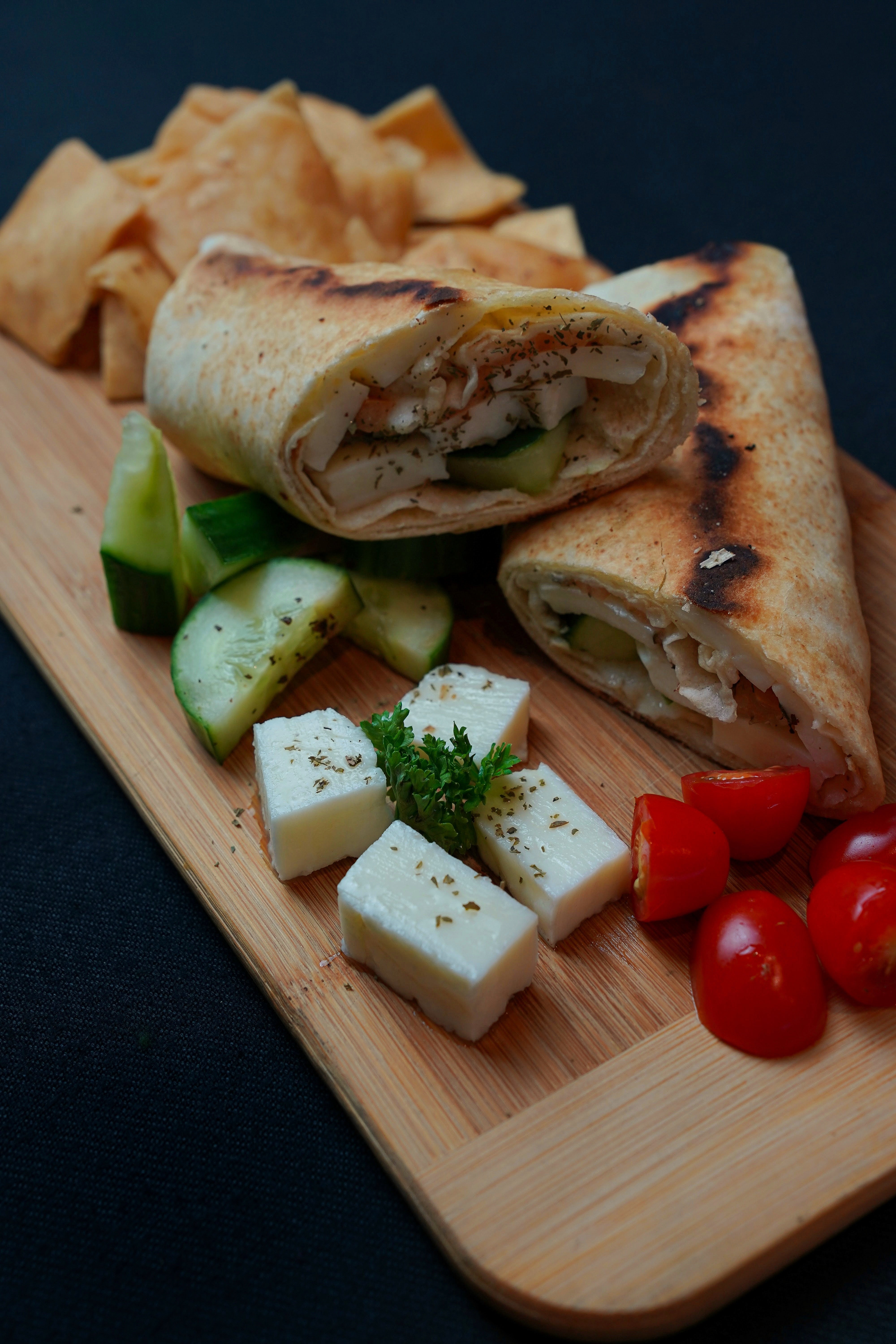 The Halloumi Wrap Combo by Tayybeh presents a wooden board showcasing a sliced pita wrap filled with chicken and cucumber. It's beautifully accompanied by cubes of feta cheese, cherry tomatoes, grilled Halloumi cheese slices, and crispy pita chips. The enticing setup invites you to indulge in the creamy hummus dip for a truly delightful Mediterranean feast.