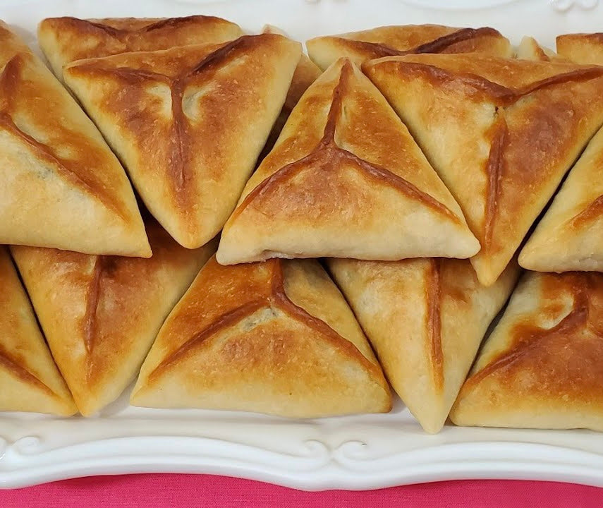 A plate of golden-brown triangular Olive Fatayer by Tayybeh, featuring hints of sweet red pepper spread, arranged neatly on a white platter.