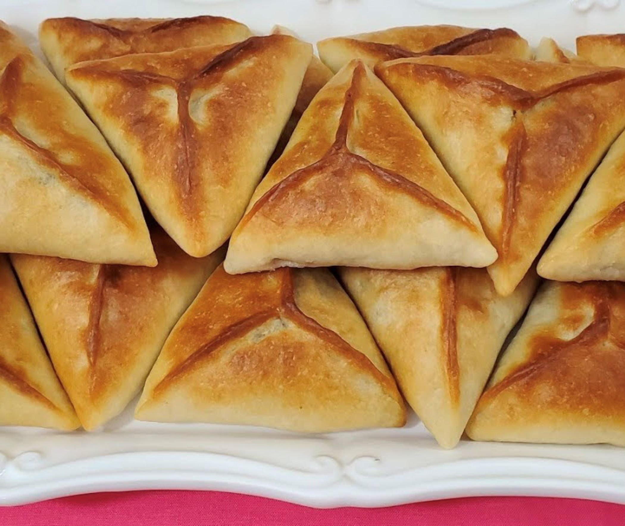 Golden-brown, triangular Tayybeh Frozen Olive Fatayer pastries, freshly baked and arranged in layers on a white platter with a pink tablecloth beneath.