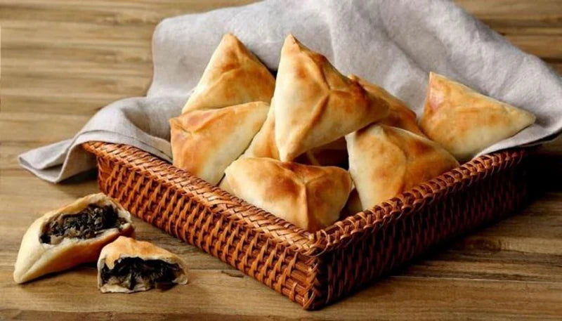 A basket of golden-brown triangular Frozen Spinach Fatayer from Tayybeh sits on a wooden table, with one fatayer revealing a rich, dark filling.