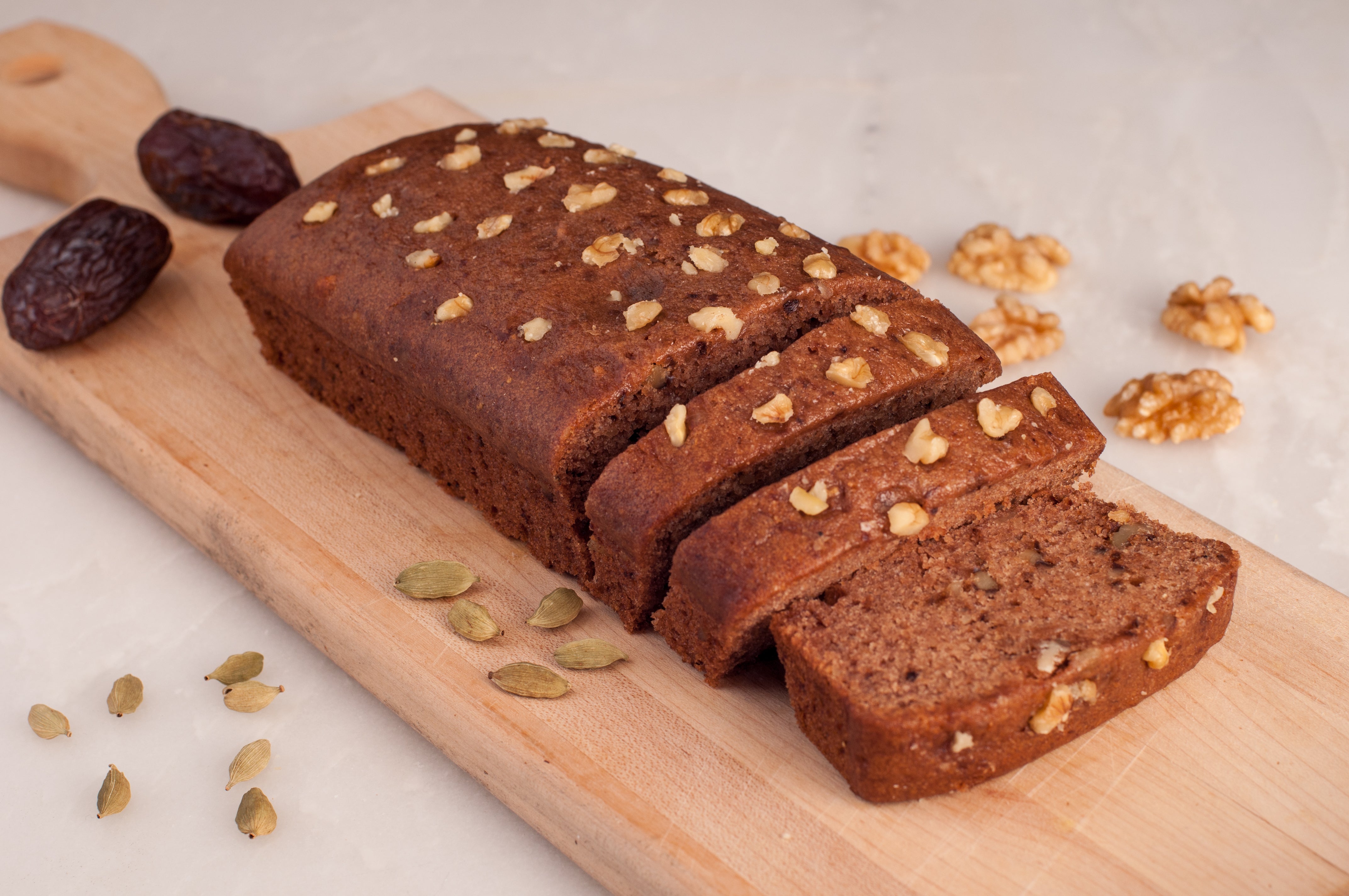 A boxed Date & Walnut Cake from Tayybeh, partially sliced and displayed on a wooden board, makes for an ideal energy snack. Ingredients such as dates, walnuts, and cardamom are clearly visible around it, emphasizing its health benefits.