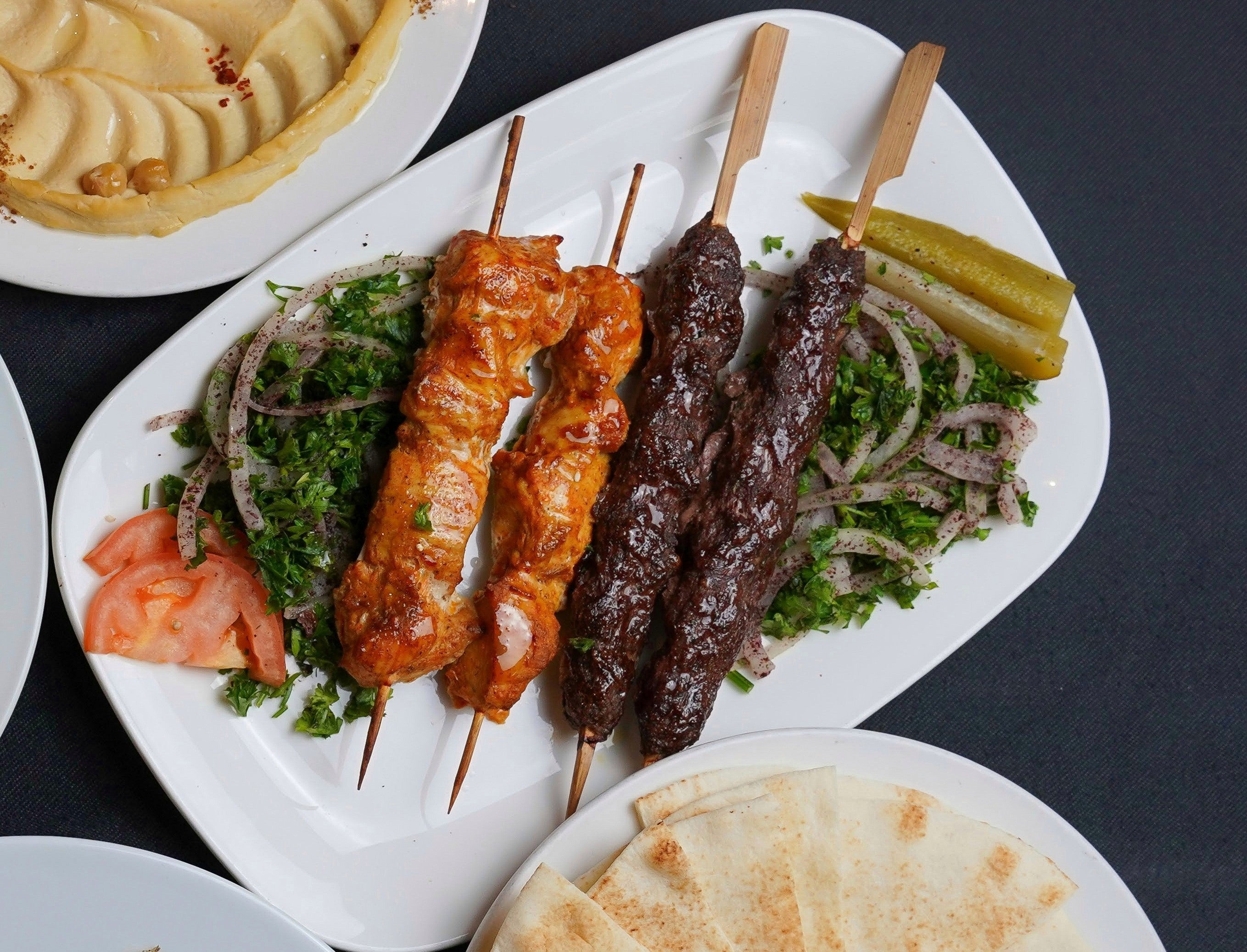 Indulge in Tayybeh's Mixed Grillz, featuring chicken Sheesh Tawook and beef kebab skewers accompanied by parsley, onions, tomato, and pickles, served with hummus and pita bread.