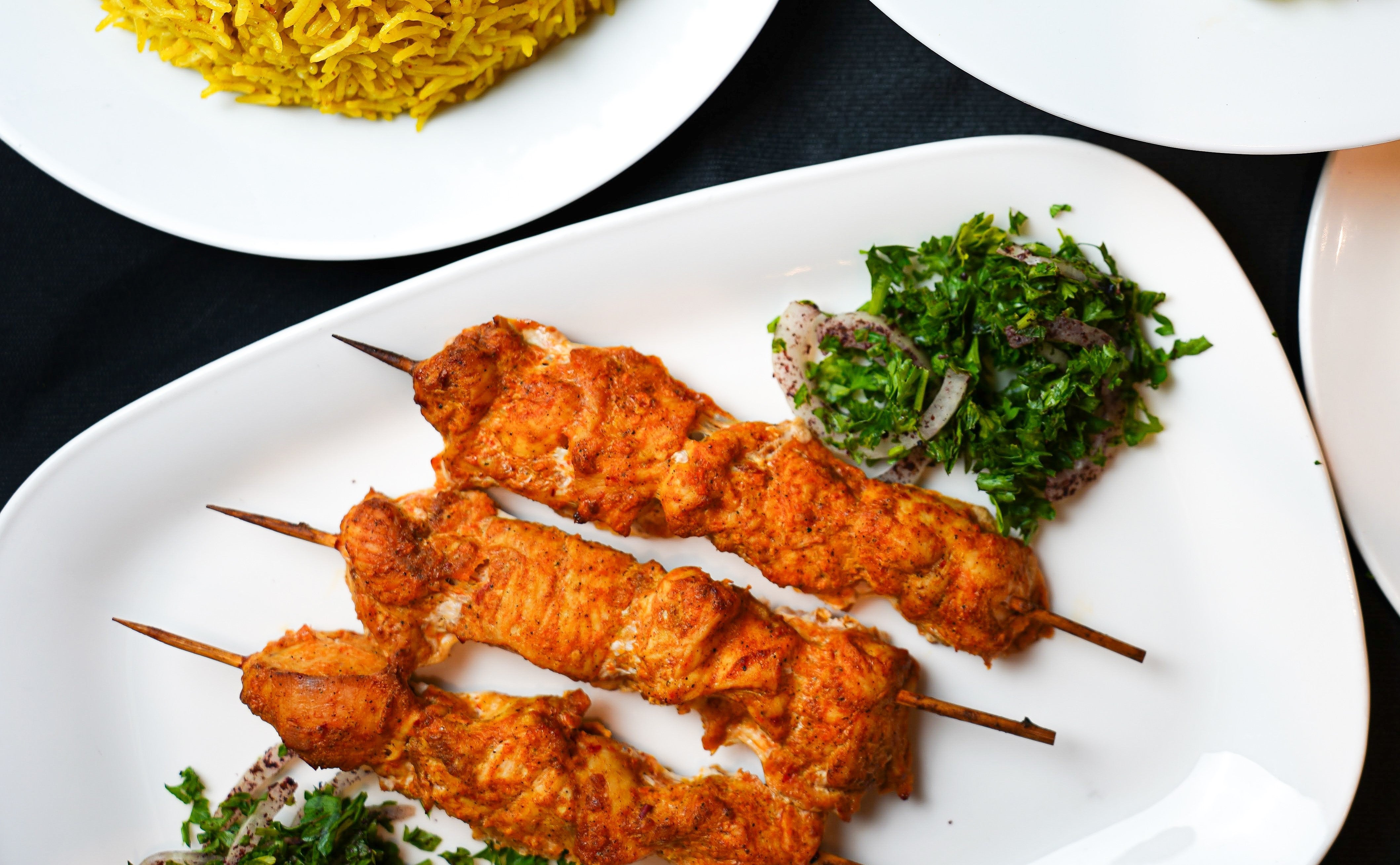 Three skewers of Tayybeh's Frozen Chicken Shish Tawook, made from tender, halal chicken breast and garnished with herbs and onions, elegantly presented on a plate beside a serving of yellow rice.