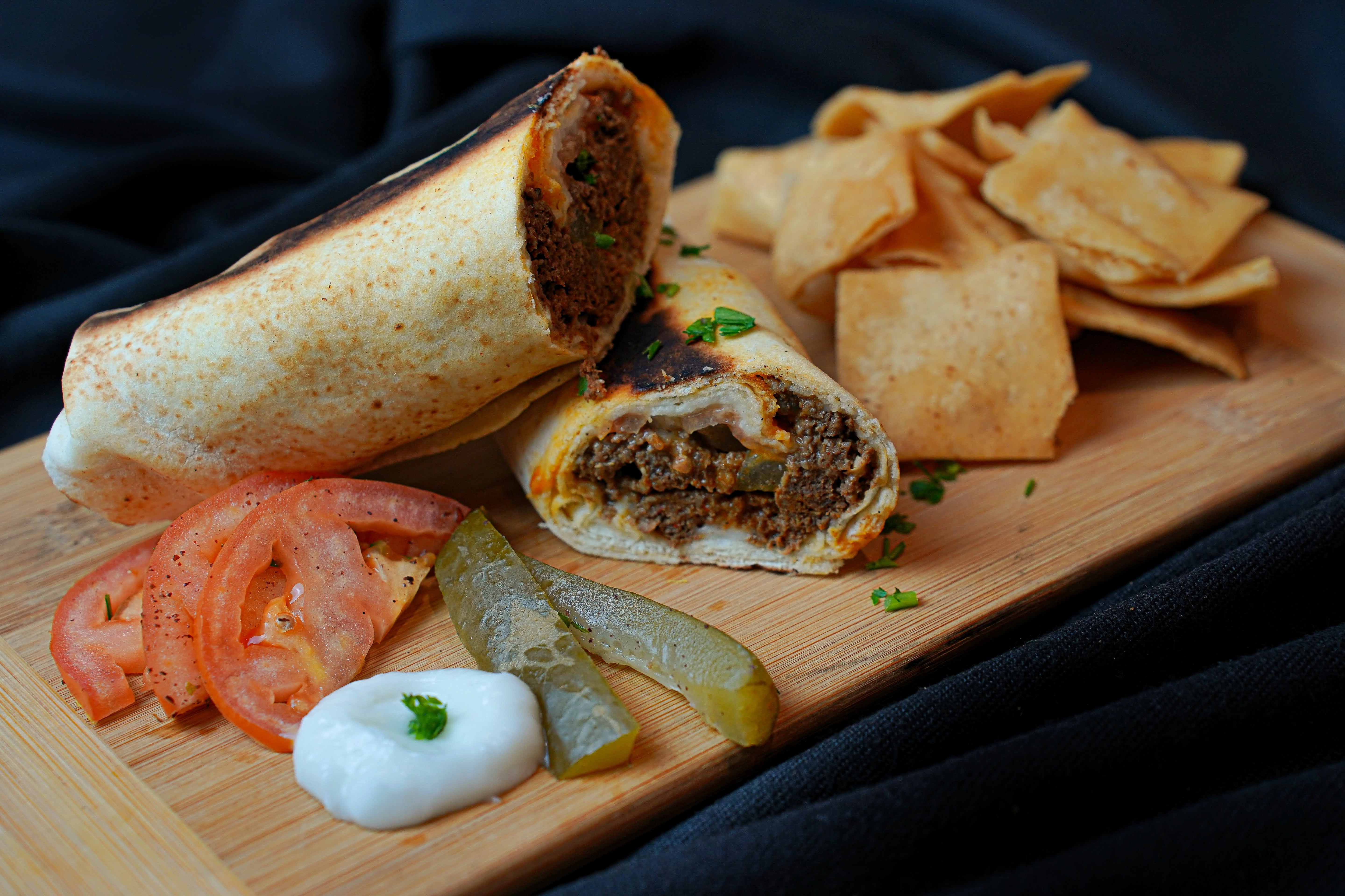 The Sujuk Wrap Combo by Tayybeh, sliced open on a wooden board, showcases its diced meat filling. It's served with tortilla chips, tomato slices, pickle slices, and a dollop of sour cream. An infusion of garlic sauce adds an extra layer of deliciousness to the dish.
