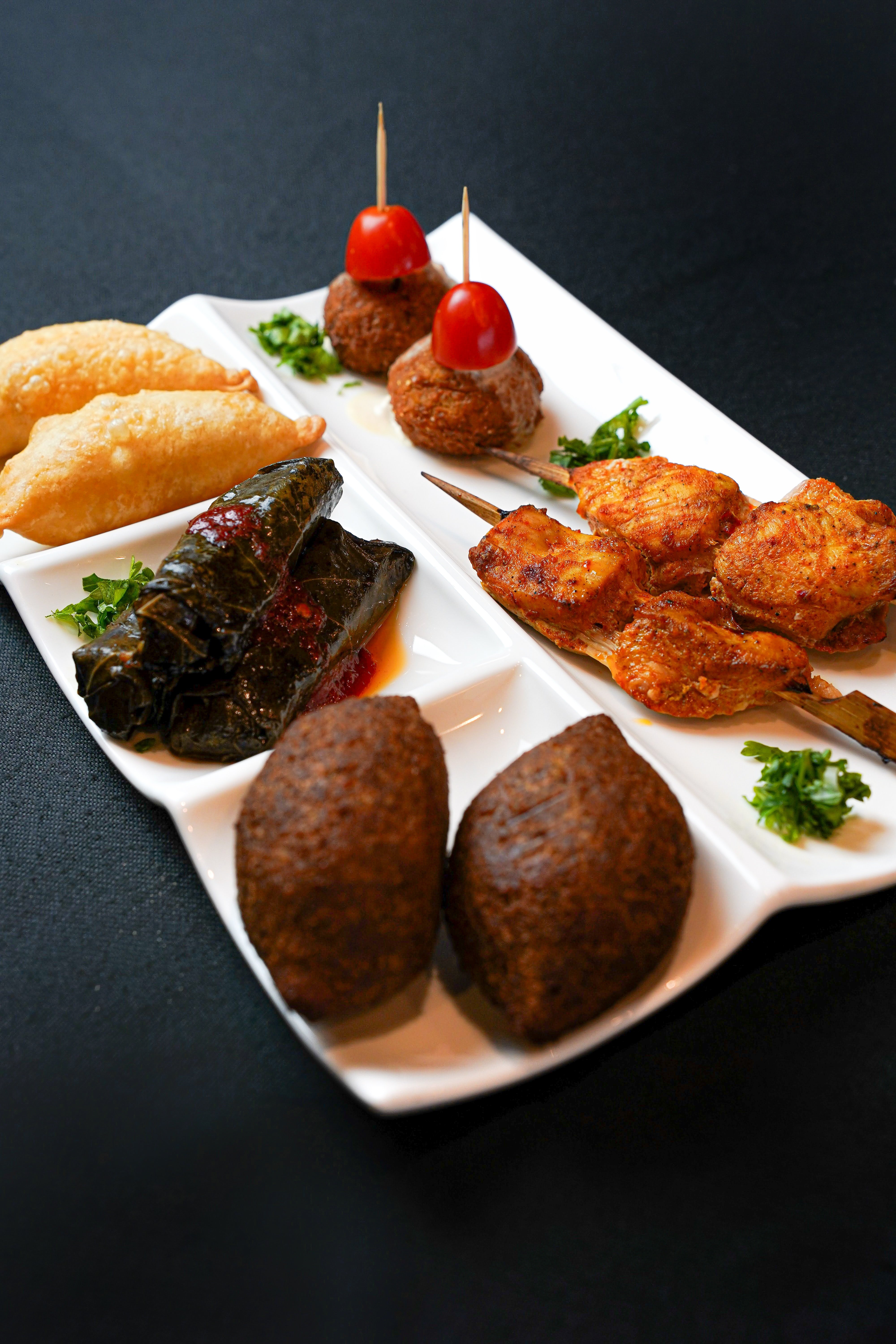 The Crowd Pleaser by Tayybeh is a plate featuring a Middle Eastern assortment of appetizers, including stuffed grape leaves, fried pastries, skewered meat, and balls topped with cherry tomatoes, all beautifully garnished with parsley.