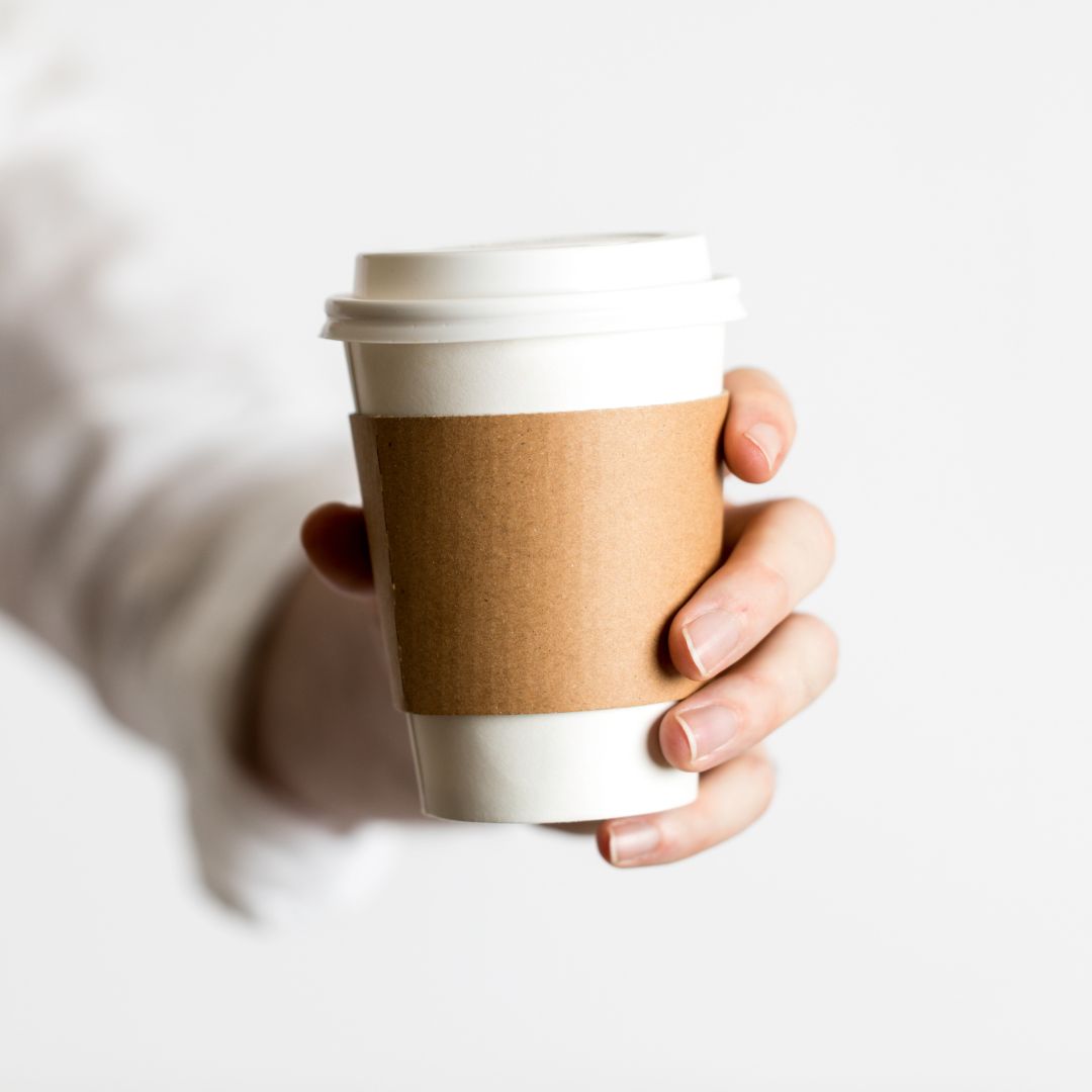A hand holds a white paper coffee cup with a brown sleeve, filled with Tayybeh's Add Coffee & Tea Service fair-trade coffee, against a plain background.