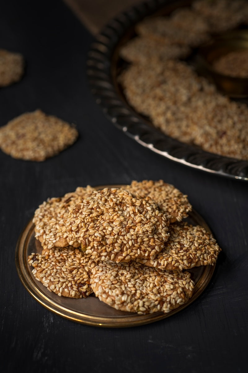 A plate of Tayybeh's Barazek is placed on a dark surface. Nearby, more of these cookies are displayed on a larger tray, tempting with their delightful texture and enticing flavors.