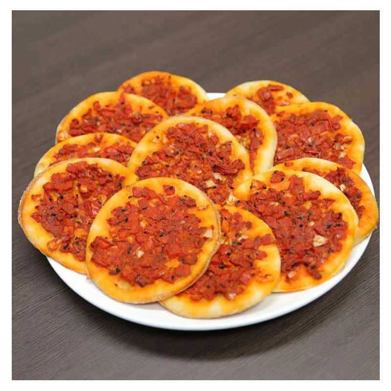 A plate of multiple round, vegan Tayybeh's Frozen Muhammara Mana'eesh, topped with minced tomatoes and herbs, is placed on a wooden table.