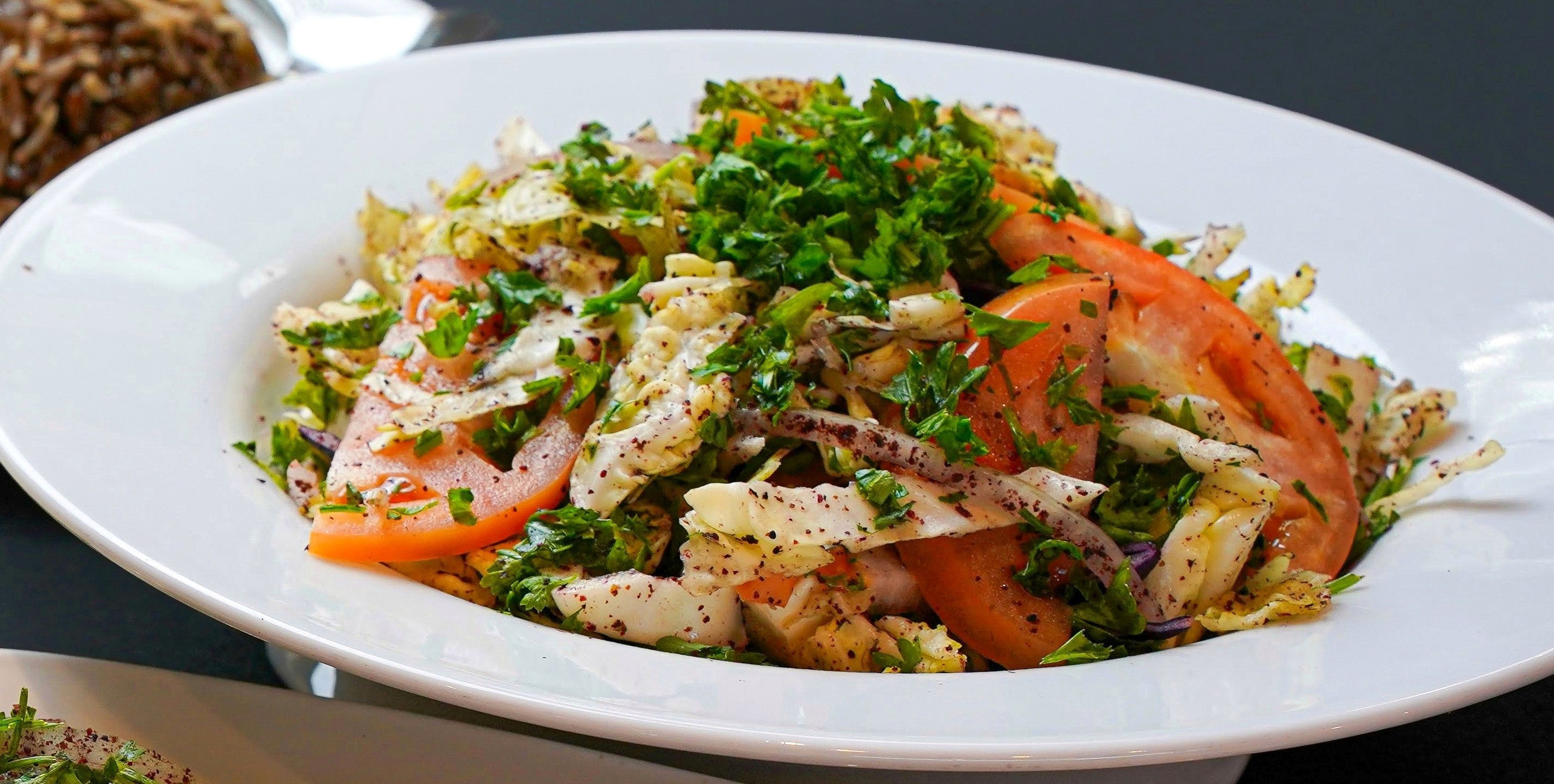 A light Malfoof Salad featuring sliced tomatoes, a refreshing cabbage slaw, and a sprinkle of sumac, all served on a white plate from the Tayybeh brand.
