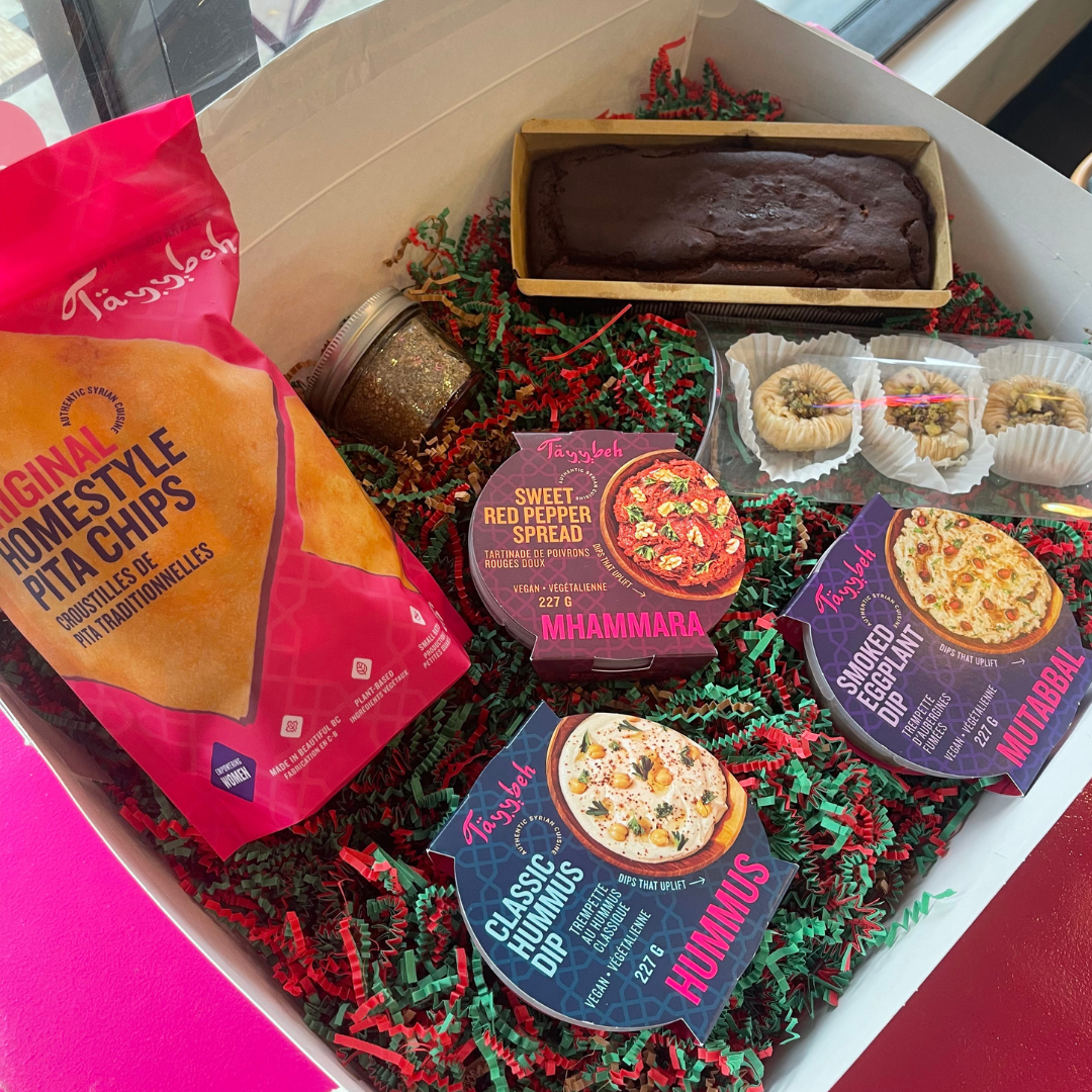 Tayybeh Premium by Tayybeh features a gift box containing original pita chips, a selection of three dip spreads (muhammara, hummus, and eggplant), a mini cake, two stuffed pastries, and a small zaatar jar, all elegantly arranged in a decorative basket.