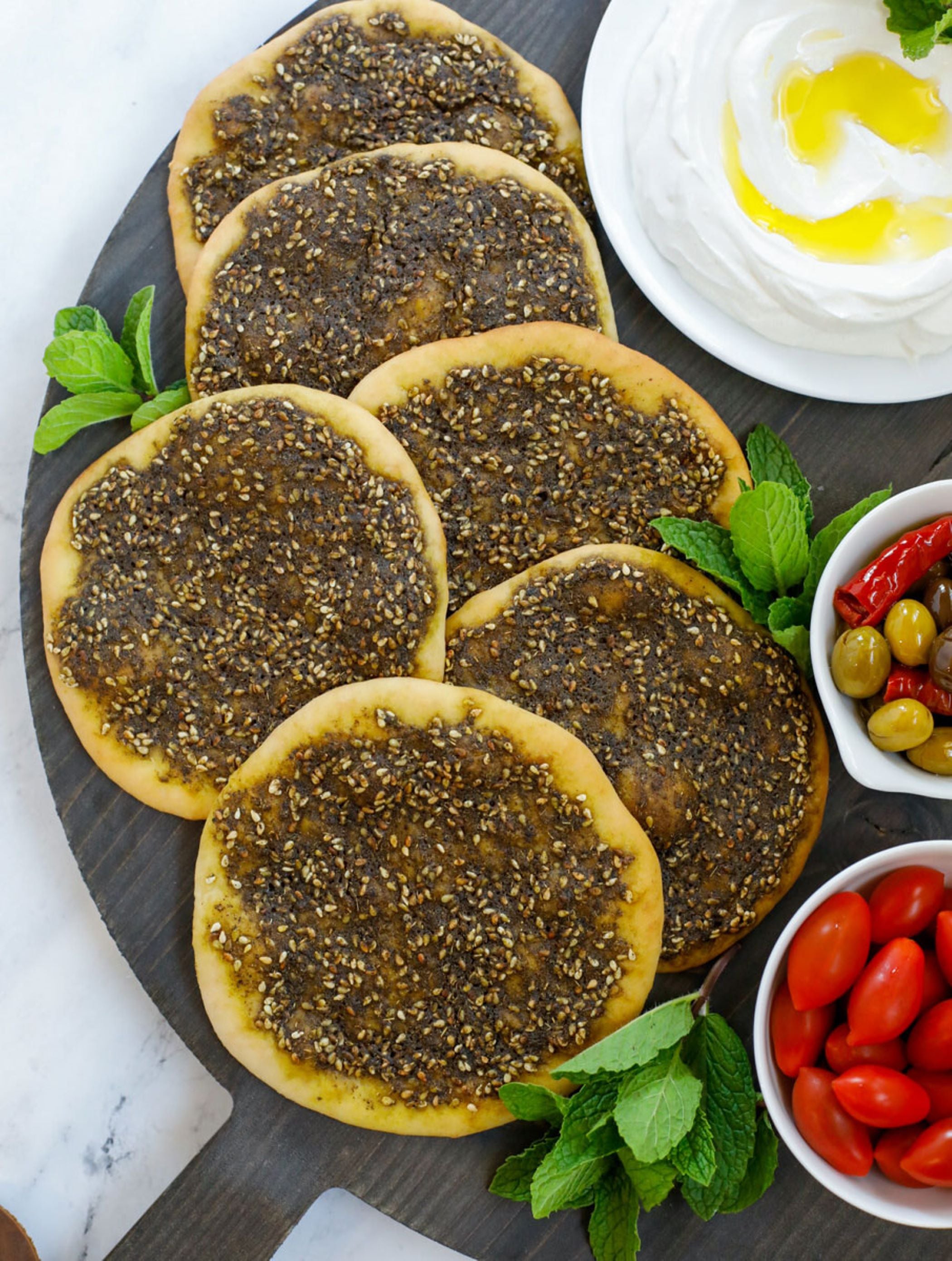 The Frozen Mana'eesh Zaatar by Tayybeh, six pieces in total, are displayed on a wooden board. They are accompanied by bowls of olives, cherry tomatoes, and yogurt drizzled with olive oil. Fresh mint leaves beautifully garnish this vegan flatbread feast.