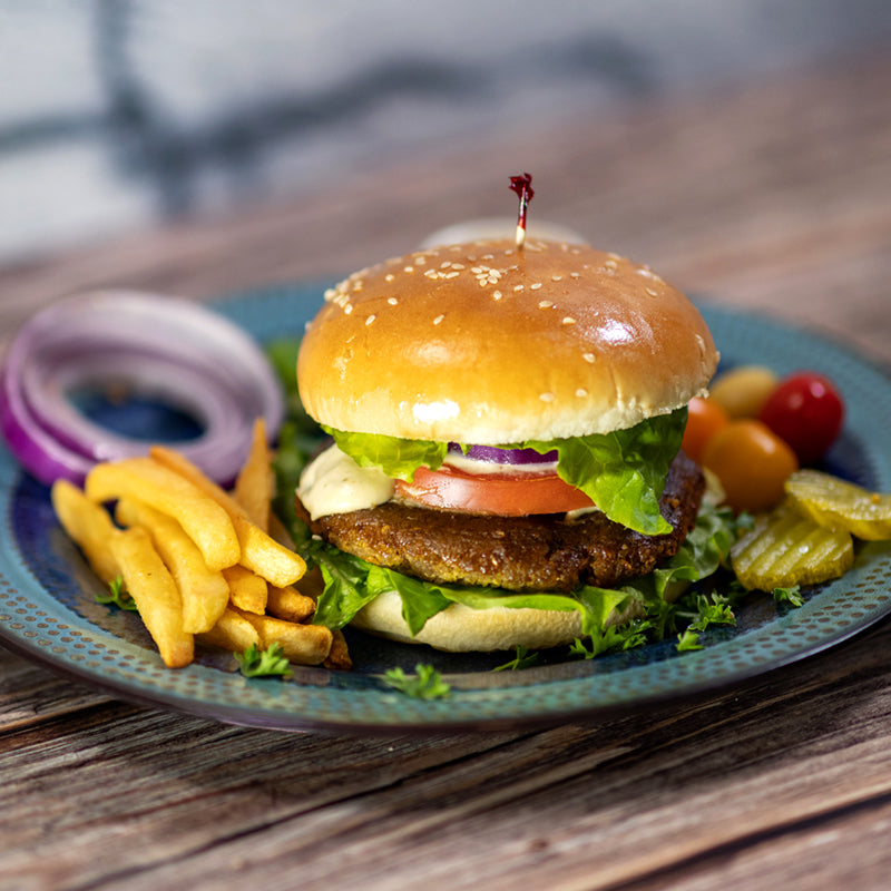 Indulge in the scrumptious Frozen Falafel Burgers by Tayybeh, featuring chickpea patties nestled between a bun and garnished with lettuce, tomato, and onion. Accompanied by crispy fries, pickles, and cherry tomatoes, this meal offers a delightful experience.