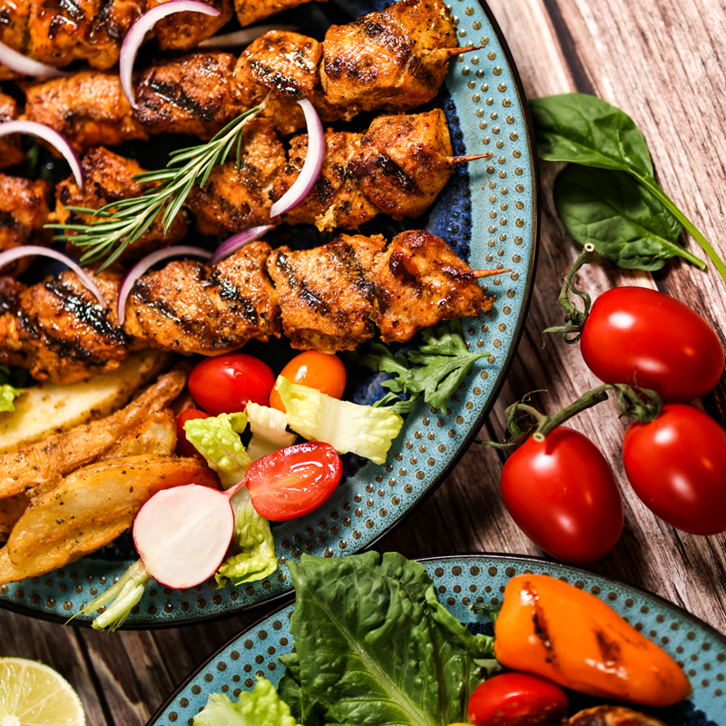 The Mini Chicken Sheesh Tawook, crafted by Tayybeh, served with rosemary, potato wedges, lettuce, cherry tomatoes, radishes, and bell peppers, creates a delightful gluten-free meal presented on a rustic wooden table.