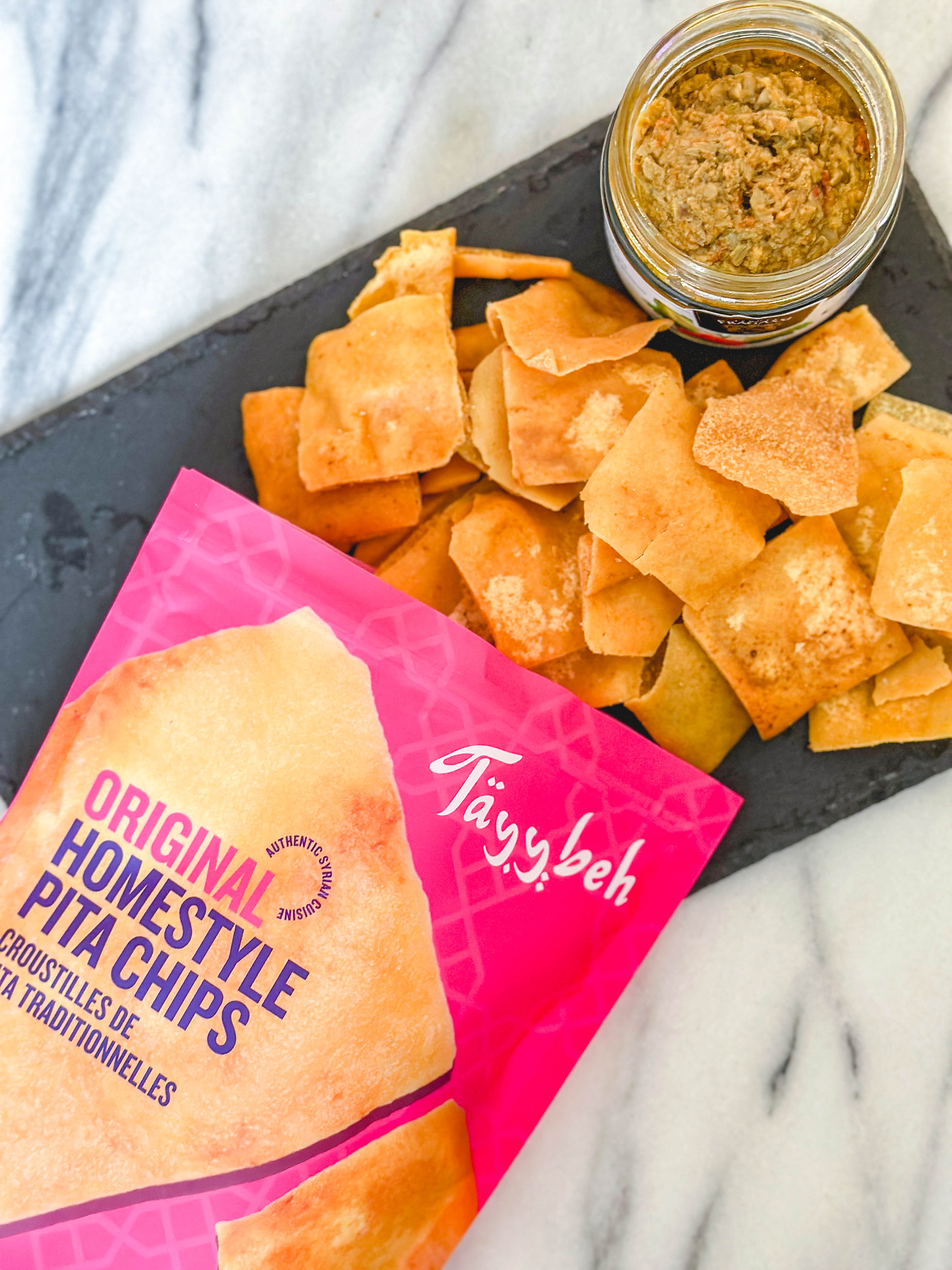 A bag of Tayybeh Homestyle Pita Chips rests beside a jar of dip on a dark serving board, all perfectly arranged on a marble countertop. Enjoy these vegan and nut-free delights at your next gathering.