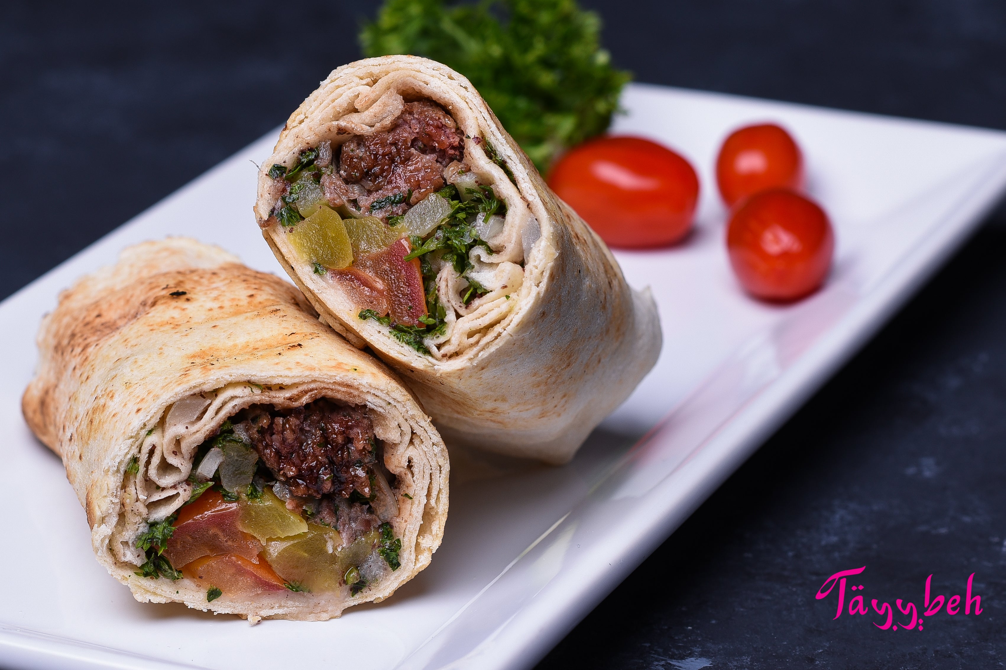 Two Tayybeh Beef Kebab Wraps, filled with savory all-beef kebab, vegetables, and herbs, are sliced in half and placed on a white rectangular plate. A creamy tahini sauce complements the dish, while cherry tomatoes and parsley garnish the background.