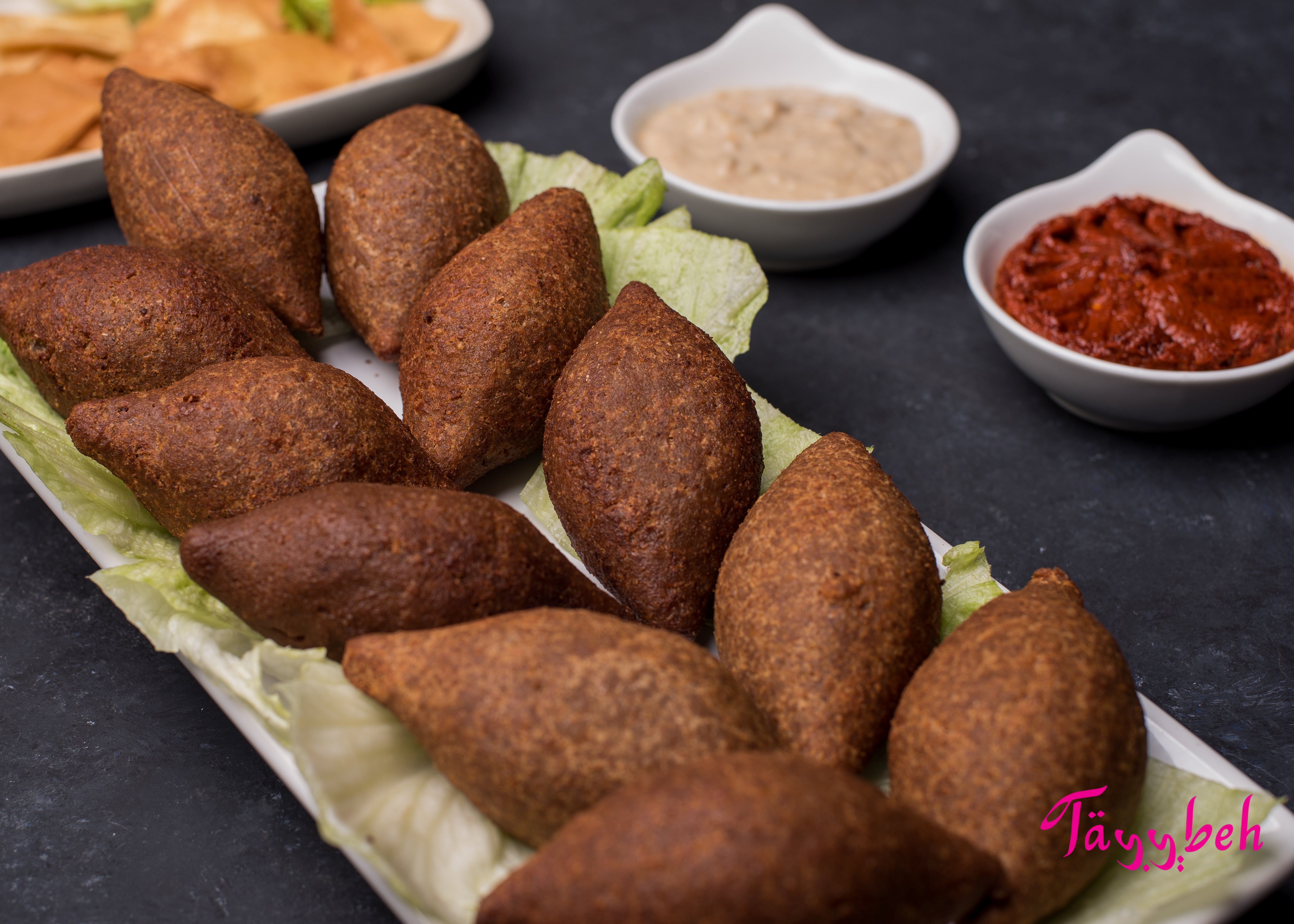 A plate of Tayybeh's Kibbeh is beautifully arranged on crisp lettuce, accompanied by small dishes of hummus and red pepper dip, all infused with rich Middle Eastern spices.