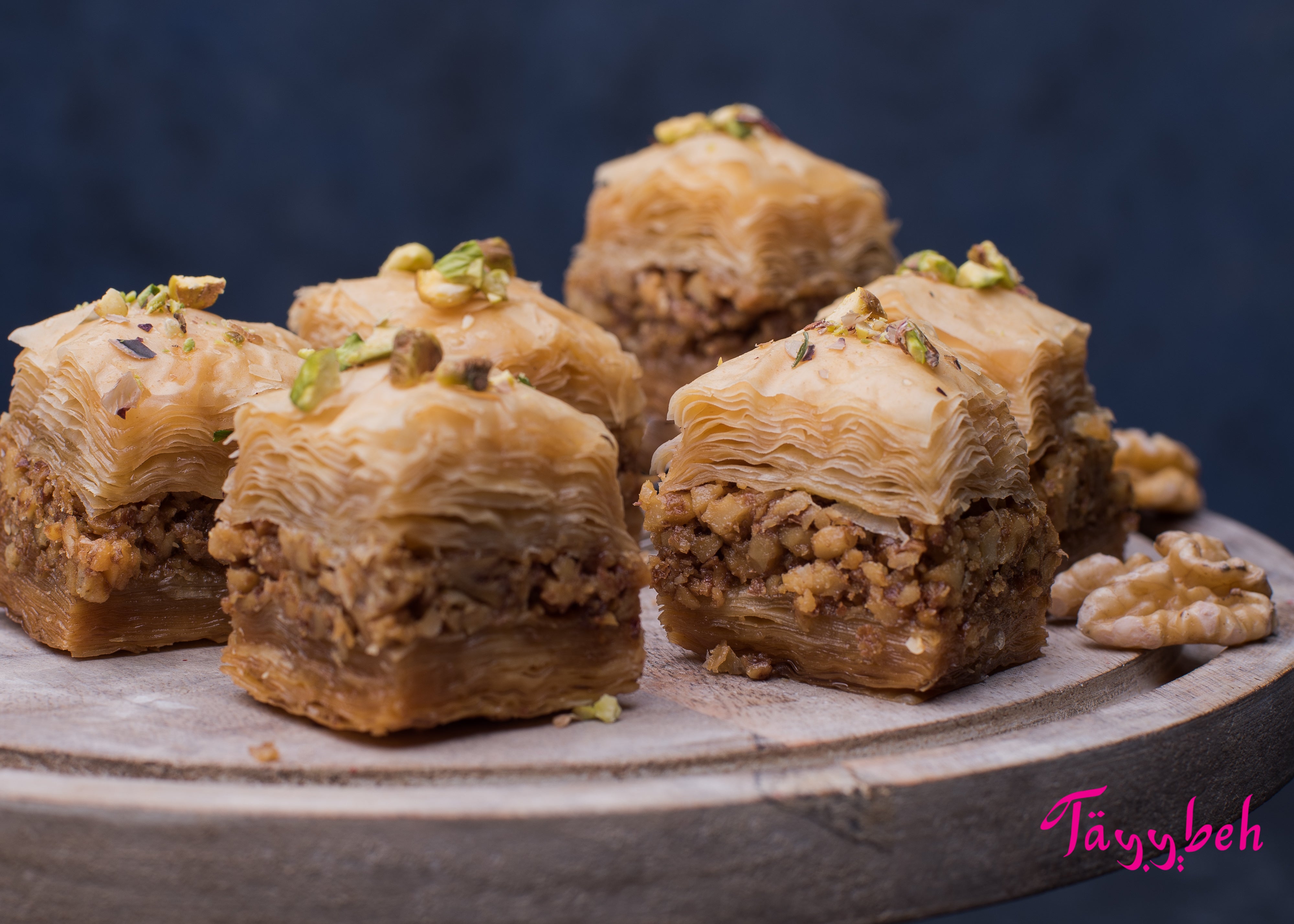 Close-up of six pieces of Tayybeh Baklava showcasing visible layers of phyllo pastry and chopped walnuts on a wooden platter, drizzled with sweet syrup and garnished with crushed pistachios.