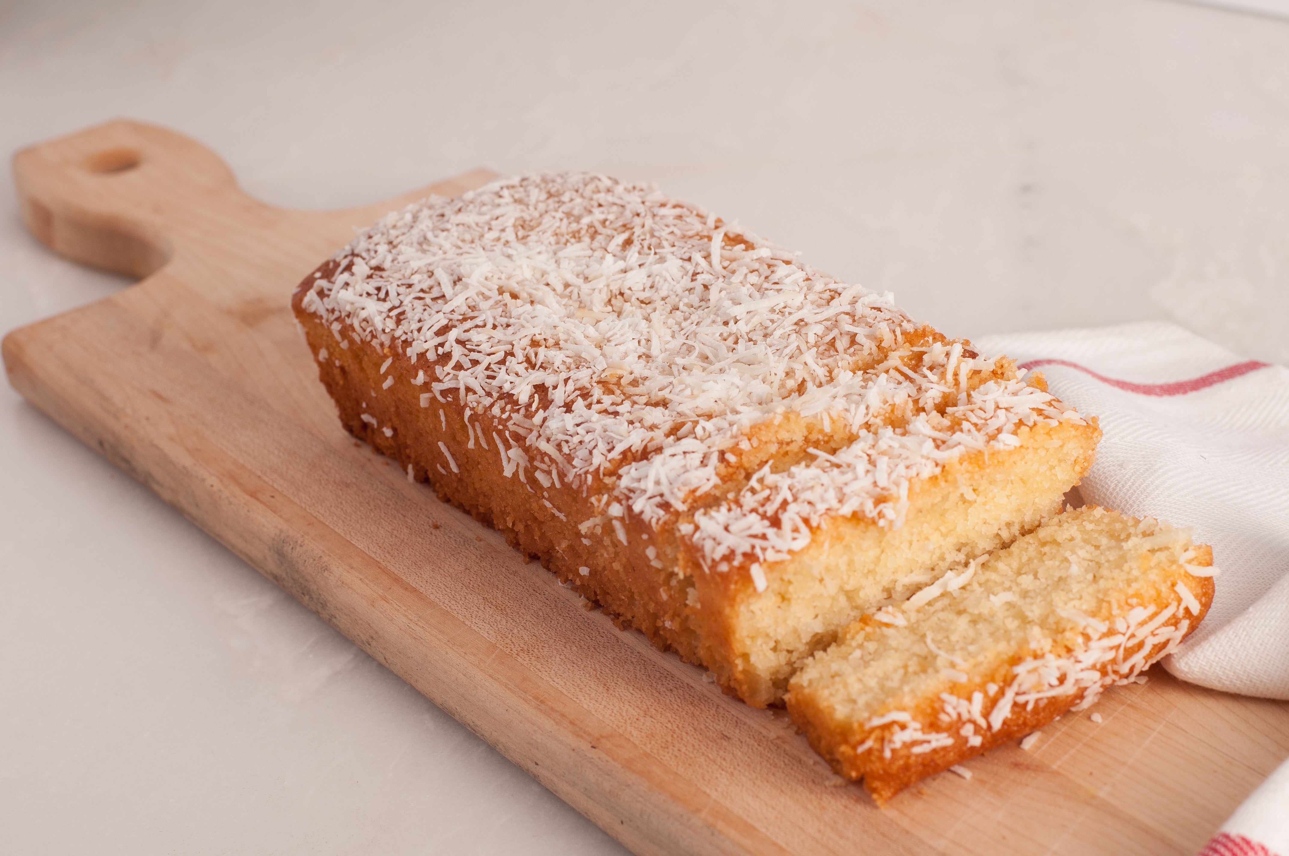 A Nammoura Loaf, a rectangular cake by Tayybeh adorned with shredded coconut topping, is placed on a wooden cutting board. A slice of this delightful vegetarian dessert is cut and partially removed. A white and red cloth in the background adds a touch of charm to this nut-free treat.