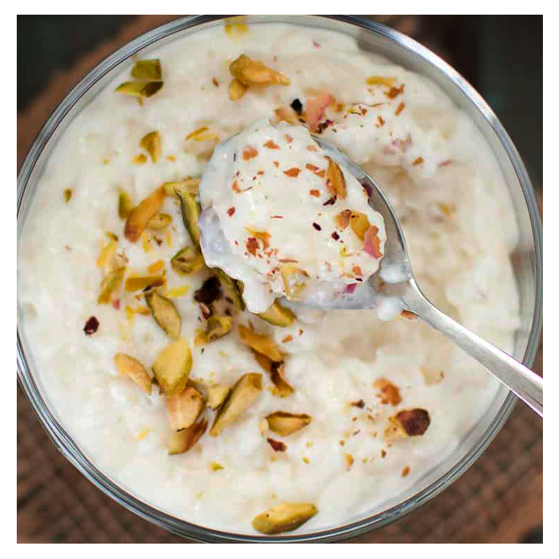 A Tayybeh Rice Pudding presented in a glass bowl, infused with fragrant rosewater, and garnished with crushed pistachios and almonds. A spoon lifts a portion, unveiling the creamy texture of the dessert.