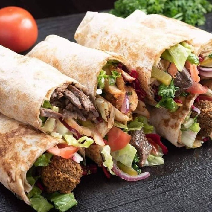 Three vegetarian wraps from the Assorted Vegetarian Wrap Platter by Tayybeh, filled with colorful ingredients and flavor, are elegantly arranged on a dark surface, with a vibrant tomato and fresh parsley adding to the background ambiance.