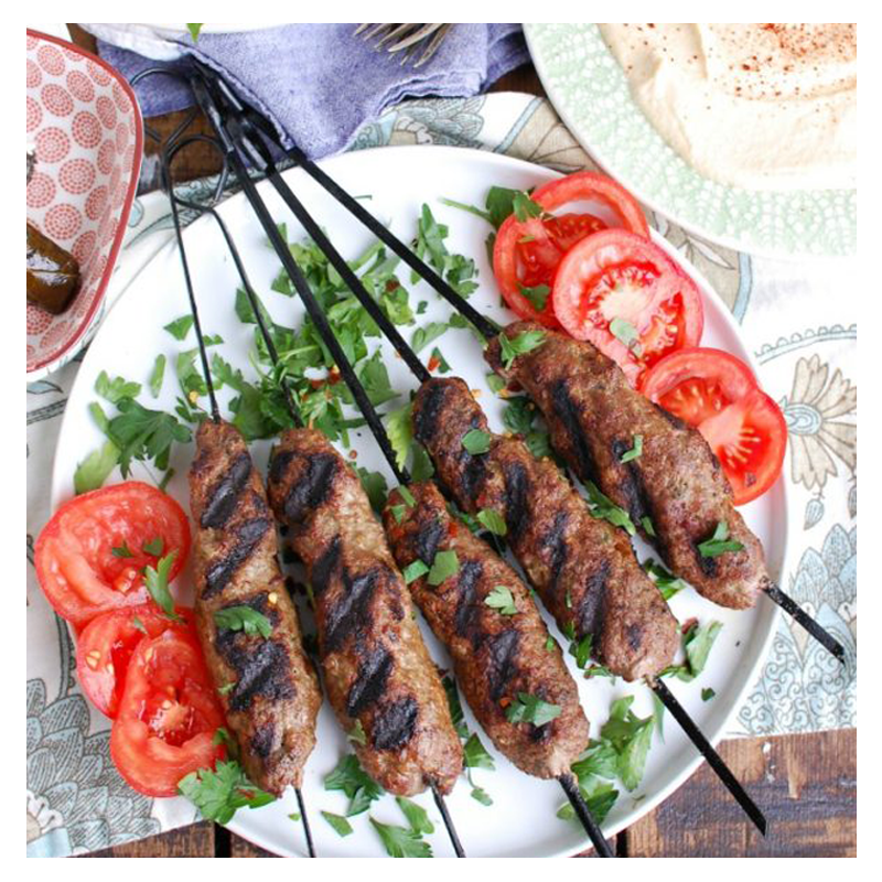 The Aleppo by Tayybeh features grilled beef kebabs with char marks, garnished with parsley, and served with creamy hummus on a white plate adorned with sliced tomatoes.