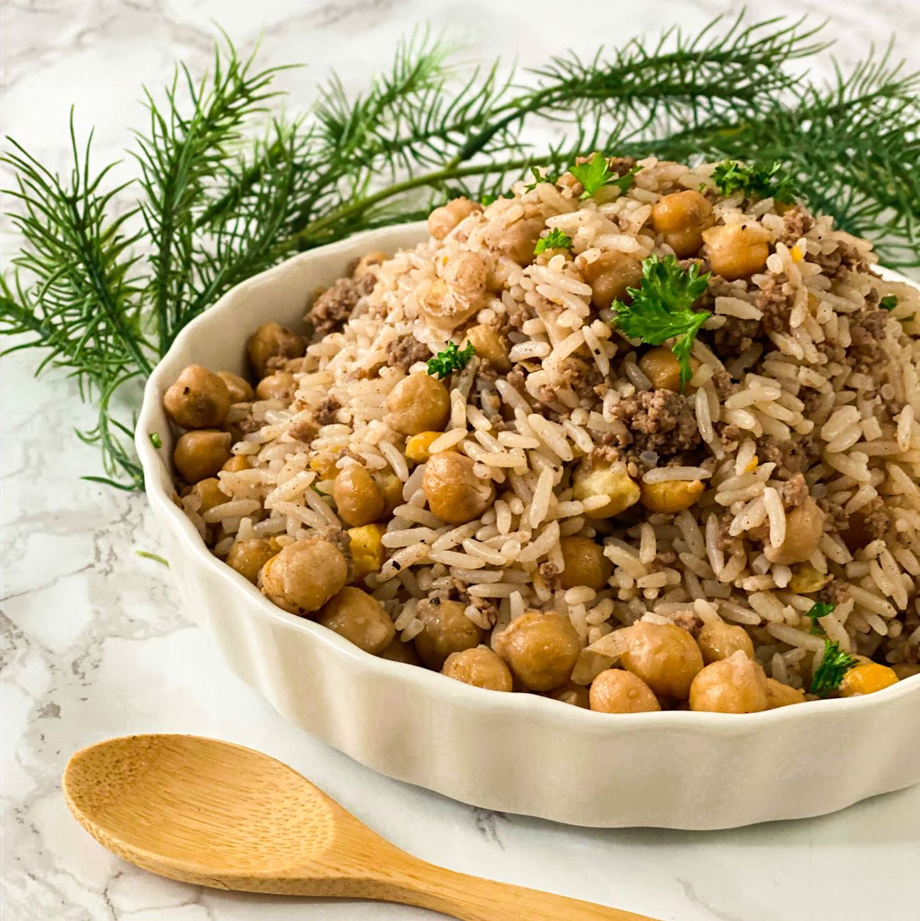 A bowl of Tayybeh's Chickpea Pilaf, featuring seasoned chickpeas and ground meat, garnished with herbs and aromatic spices, rests on a marble surface next to a wooden spoon and sprigs of fresh dill.