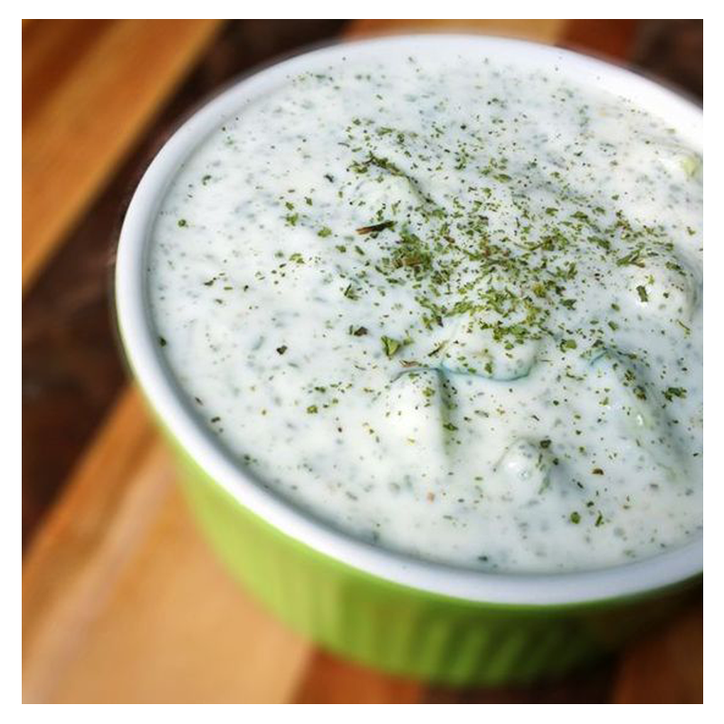 A Tayybeh Cucumber yogurt dip, a green ramekin filled with a creamy blend infused with whipped yogurt and mint, garnished with chopped herbs on top, is placed on a wooden surface.