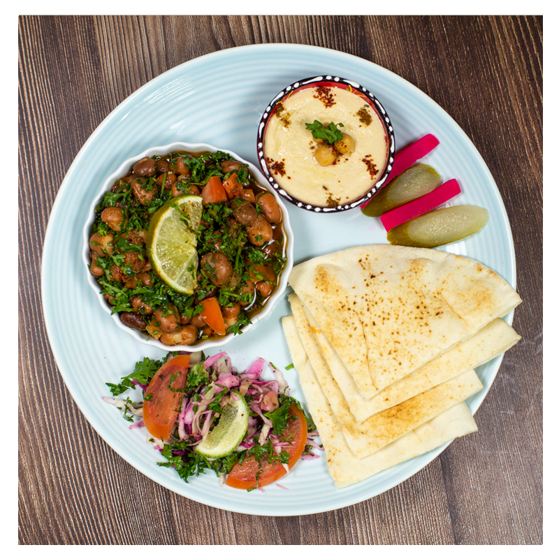 A Fava Bean Stew Meal by Tayybeh, featuring a rich hummus dip, warm pita bread, a fava bean salad with lime, pickled vegetables, and a tomato-onion salad sits invitingly on the wooden table.
