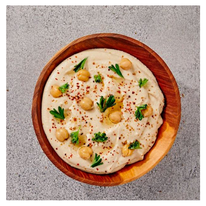 A bowl of Tayybeh's Hummus Dip, garnished with fresh chickpeas, parsley, and red pepper flakes, is placed on a gray surface.
