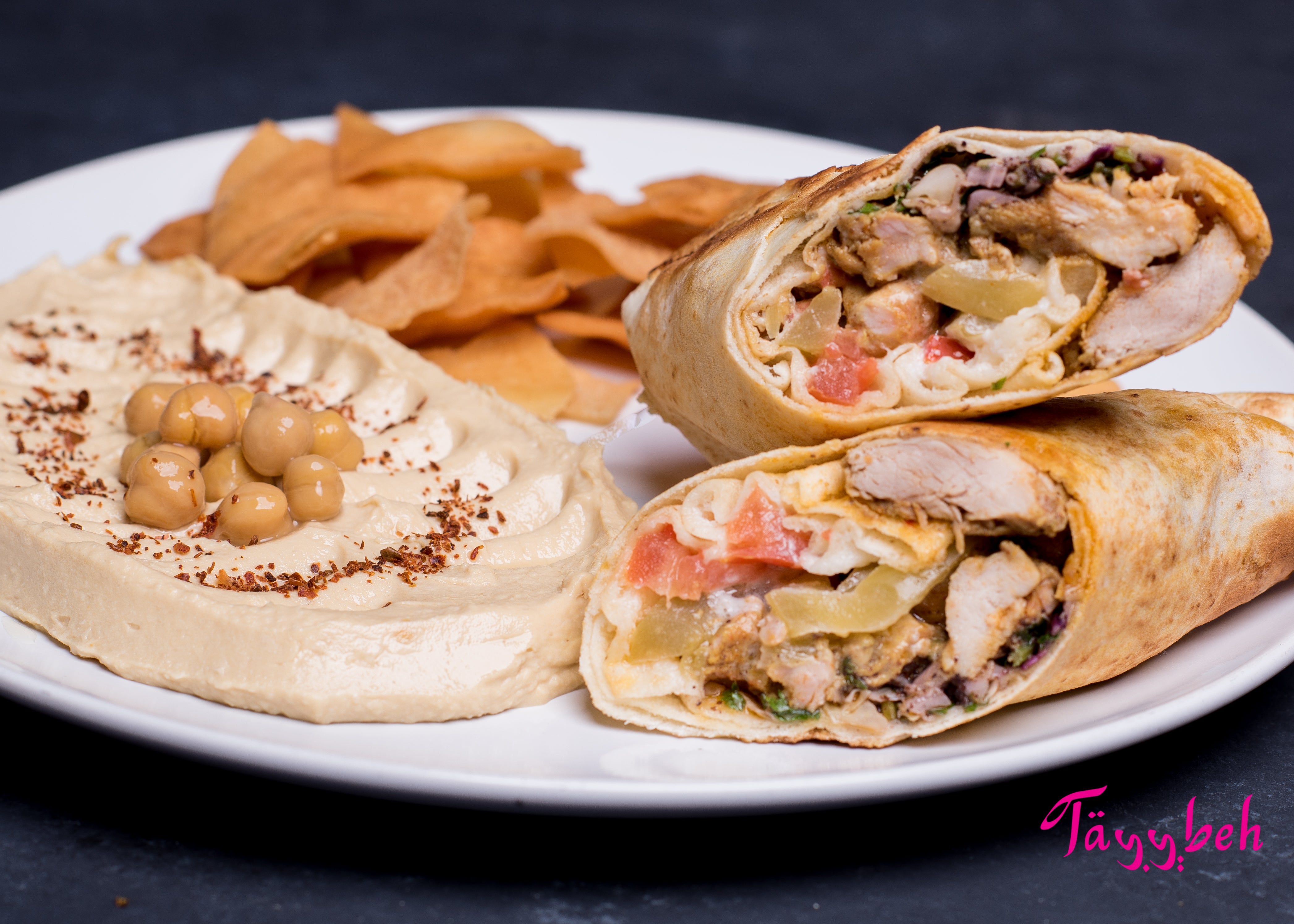 Introducing the Chicken Shawerma Wrap Combo by Tayybeh: a plate featuring a creamy hummus dip topped with chickpeas, accompanied by a sliced pita wrap stuffed with grilled chicken and vegetables, plus a side of chips.