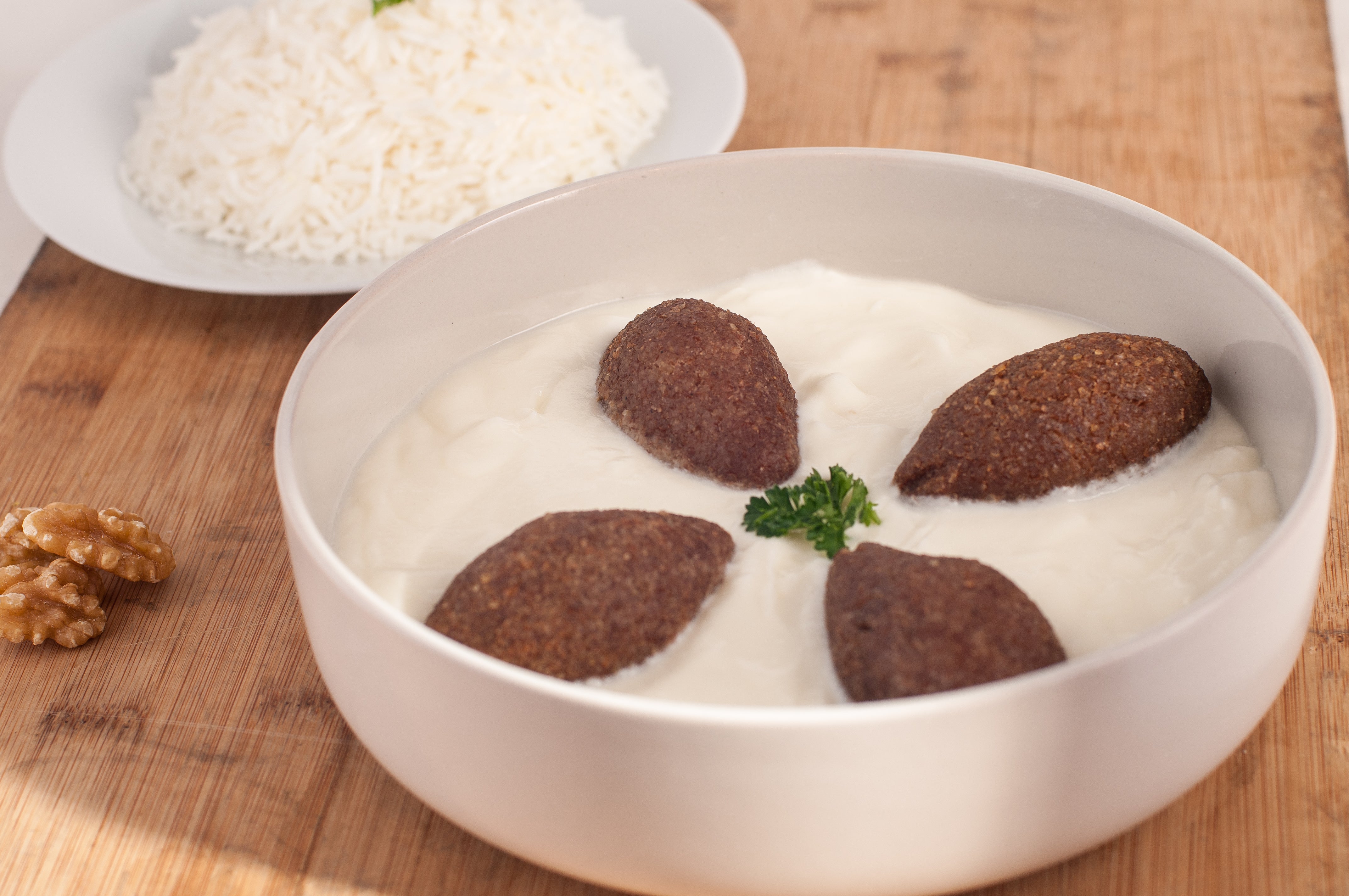 A bowl of Tayybeh’s Kibbeh be Laban, infused with Middle Eastern spices and garnished with a sprig of parsley. In the background, a plate of white rice is served alongside some walnuts.