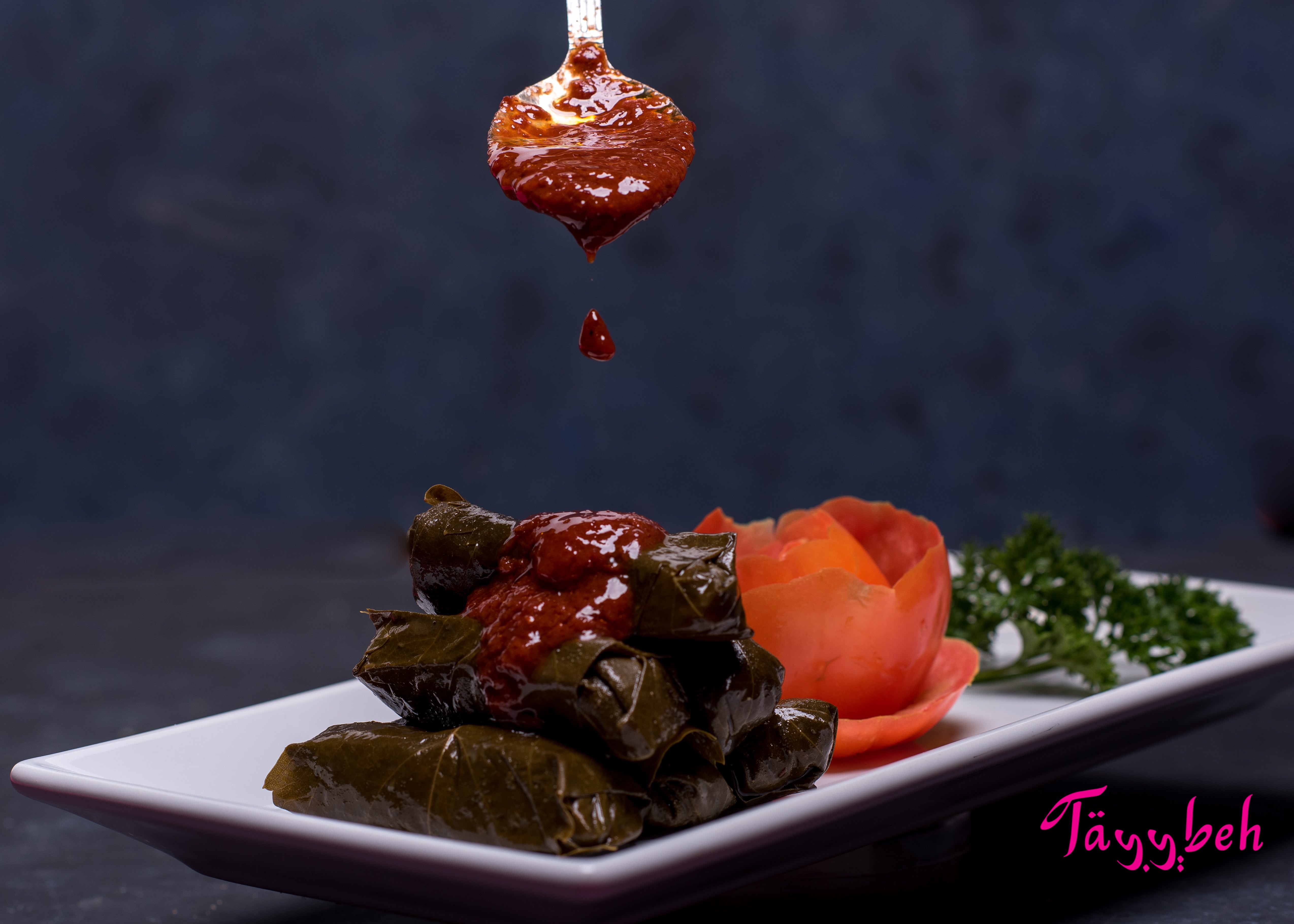 A spoon drips red sauce onto a stack of Tayybeh's Yalanji on a white plate, garnished with a tomato flower and parsley. This vegan, gluten-free delight promises to tantalize your taste buds.